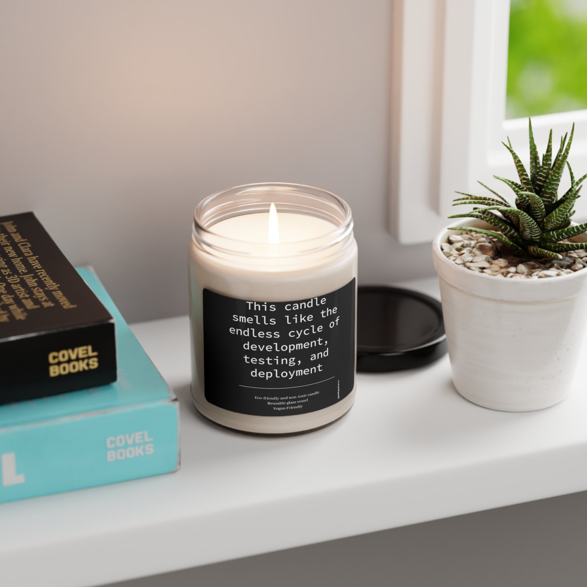 A This Candle Smells Like the Endless Cycle of Development, Testing and Deployment candle on a windowsill next to a stack of books and a potted succulent, with a humorous label about the endless cycle of development.