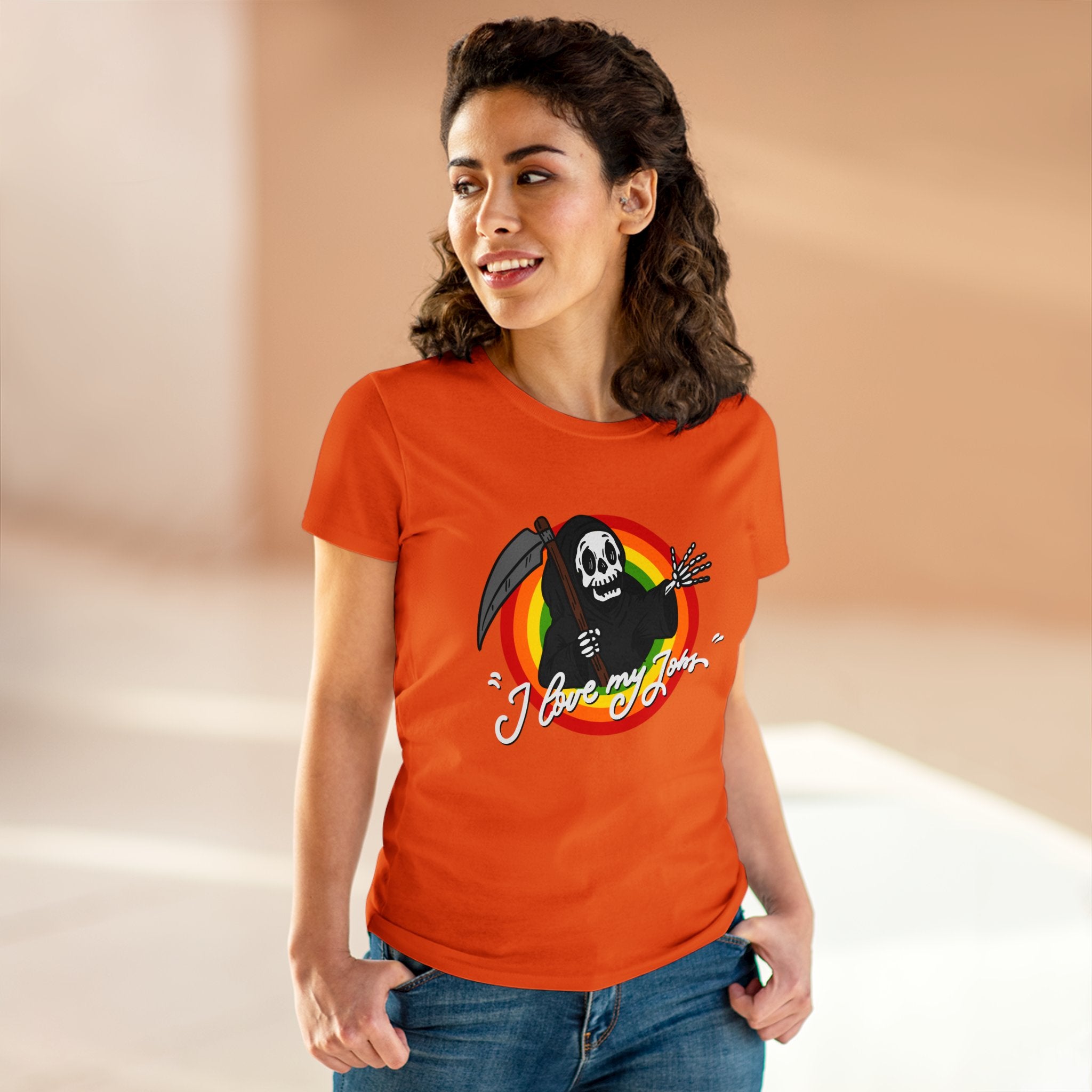 A woman with curly hair smiles and wears a soft, comfortable cotton Love My Jobs - Women's Tee, featuring a Grim Reaper graphic and the text "I Love My Job." She stands indoors against a blurred background.