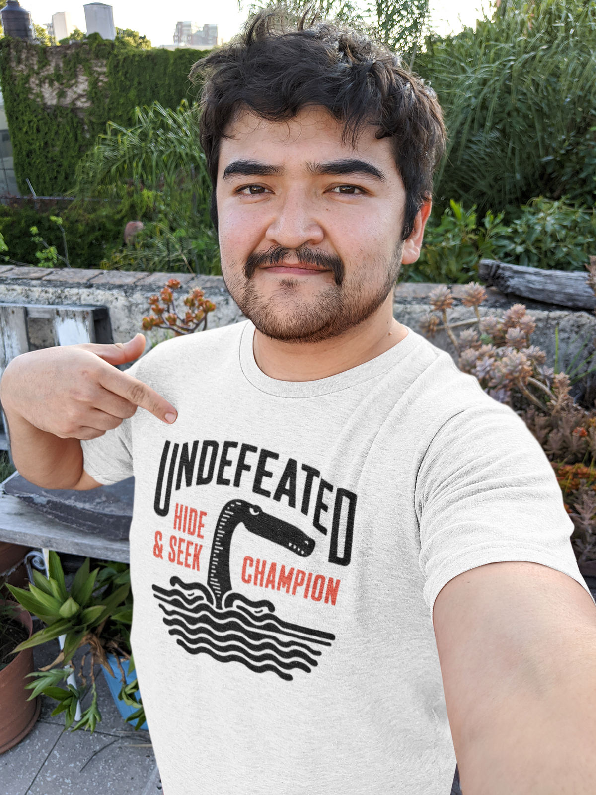 "Undefeated Hide and Seek Champion" t-shirt in white, man modeling it outdoors, funny game gift