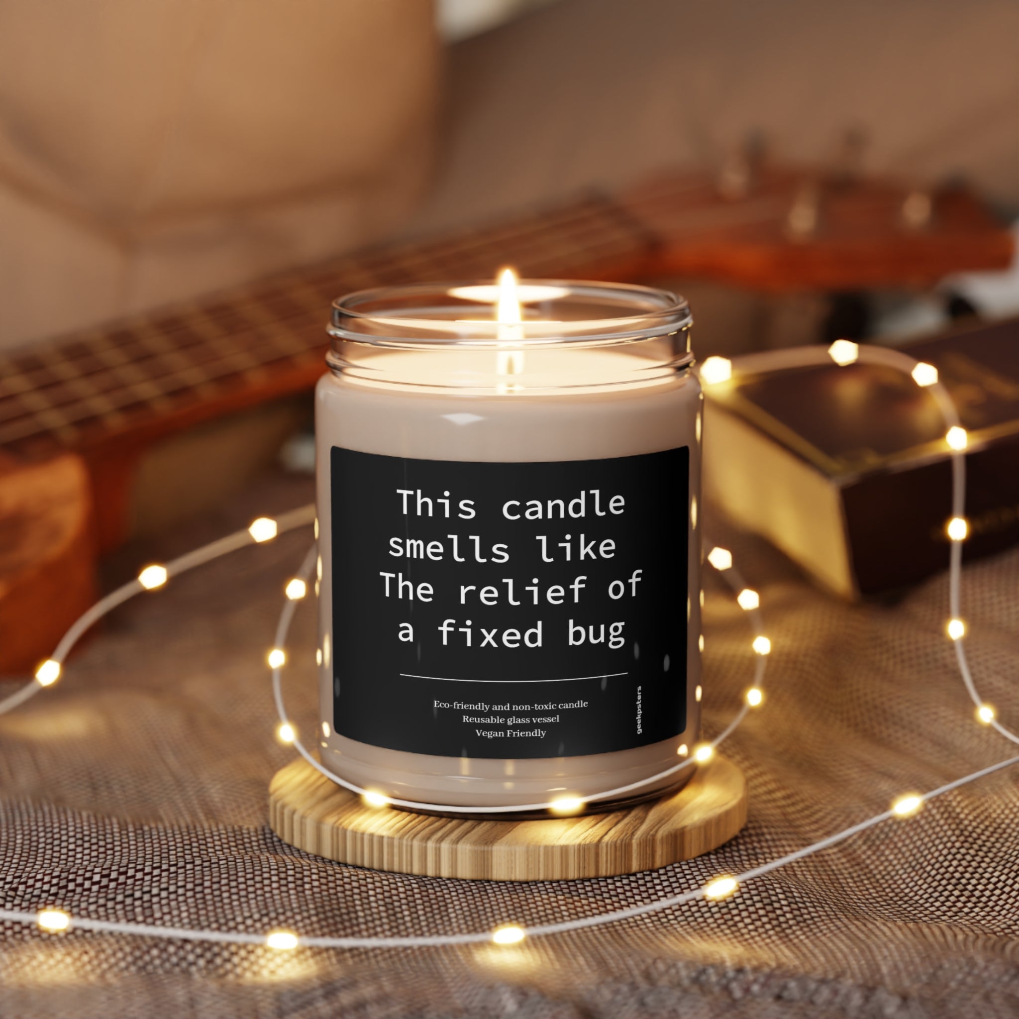 A This Candle Smells Like The Relief of a Fixed Bug scented soy candle with a label reading "this candle smells like the relief of a fixed bug," placed on a wooden coaster, surrounded by fairy lights.