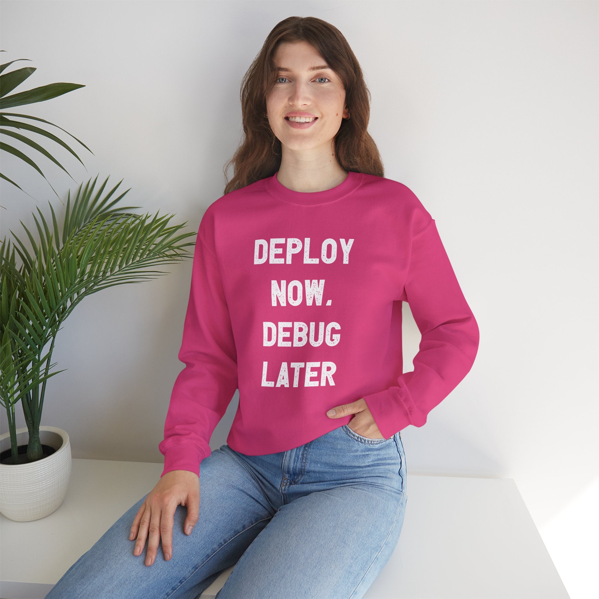 A woman in a pink "Deploy Now, Debug Later" sweatshirt sits beside a potted plant, creating the perfect cozy atmosphere for tech enthusiasts during the colder months.