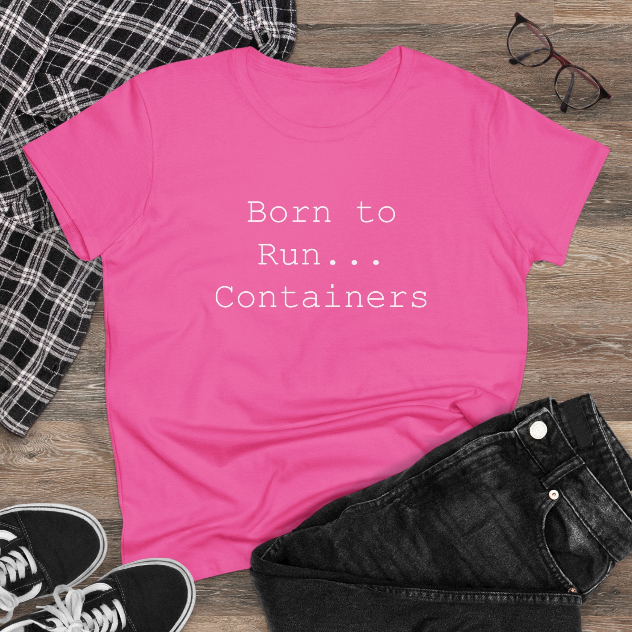 A wardrobe essential, the Born to Run Containers - Women's Tee in pink features the "Born to Run... Containers" text. It is stylishly displayed on a wooden surface with a plaid shirt, black jeans, black sneakers, and glasses.