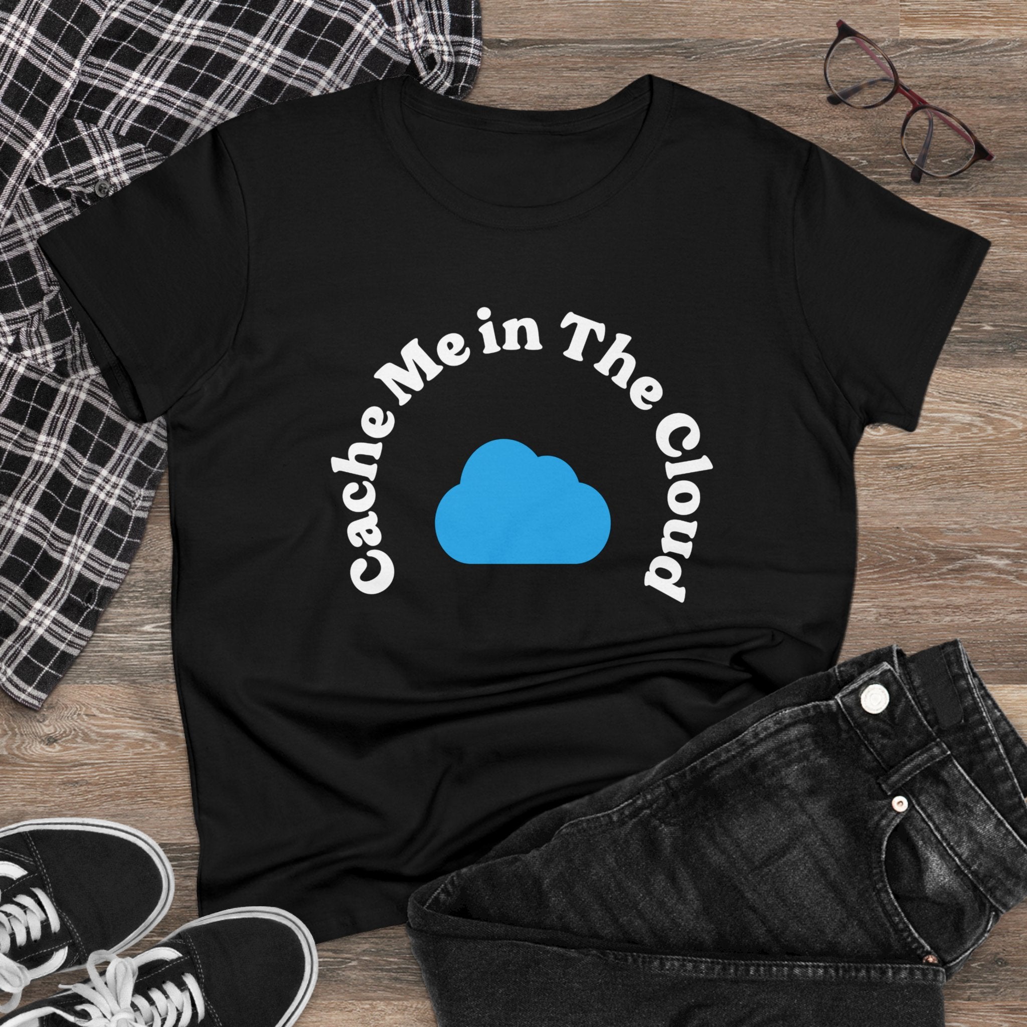 The "Cache Me in The Cloud" Women's Tee is made from light cotton and showcases a blue cloud graphic. It’s paired with black jeans, black-and-white sneakers, glasses, and a plaid shirt on a wooden surface for an effortlessly casual vibe.