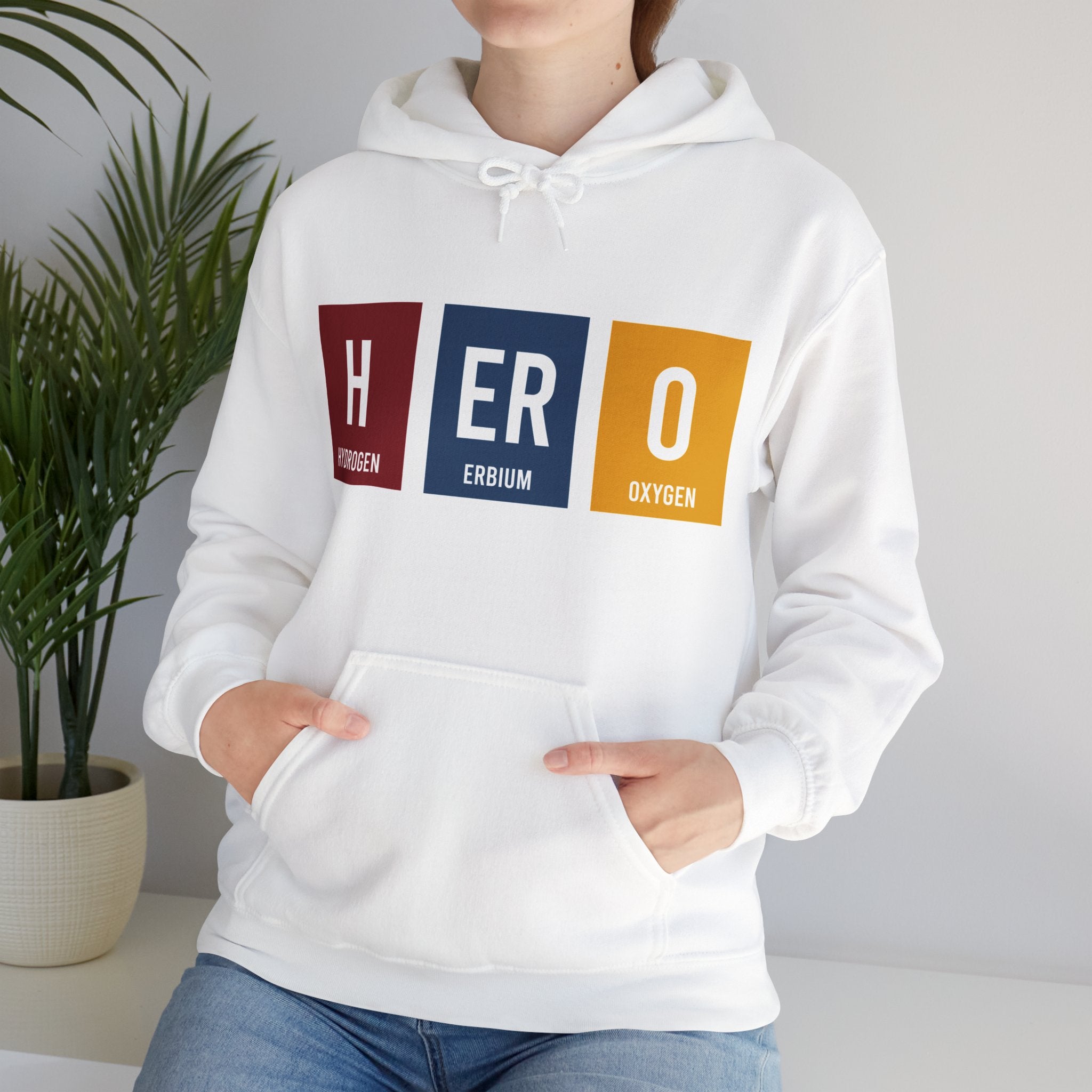 A person wearing a white **HERO - Hooded Sweatshirt** with the word "HERO" cleverly designed using the symbols for hydrogen, erbium, and oxygen from the periodic table. They are standing with hands in the hoodie’s front pocket, embodying an element of heroism in their stance.