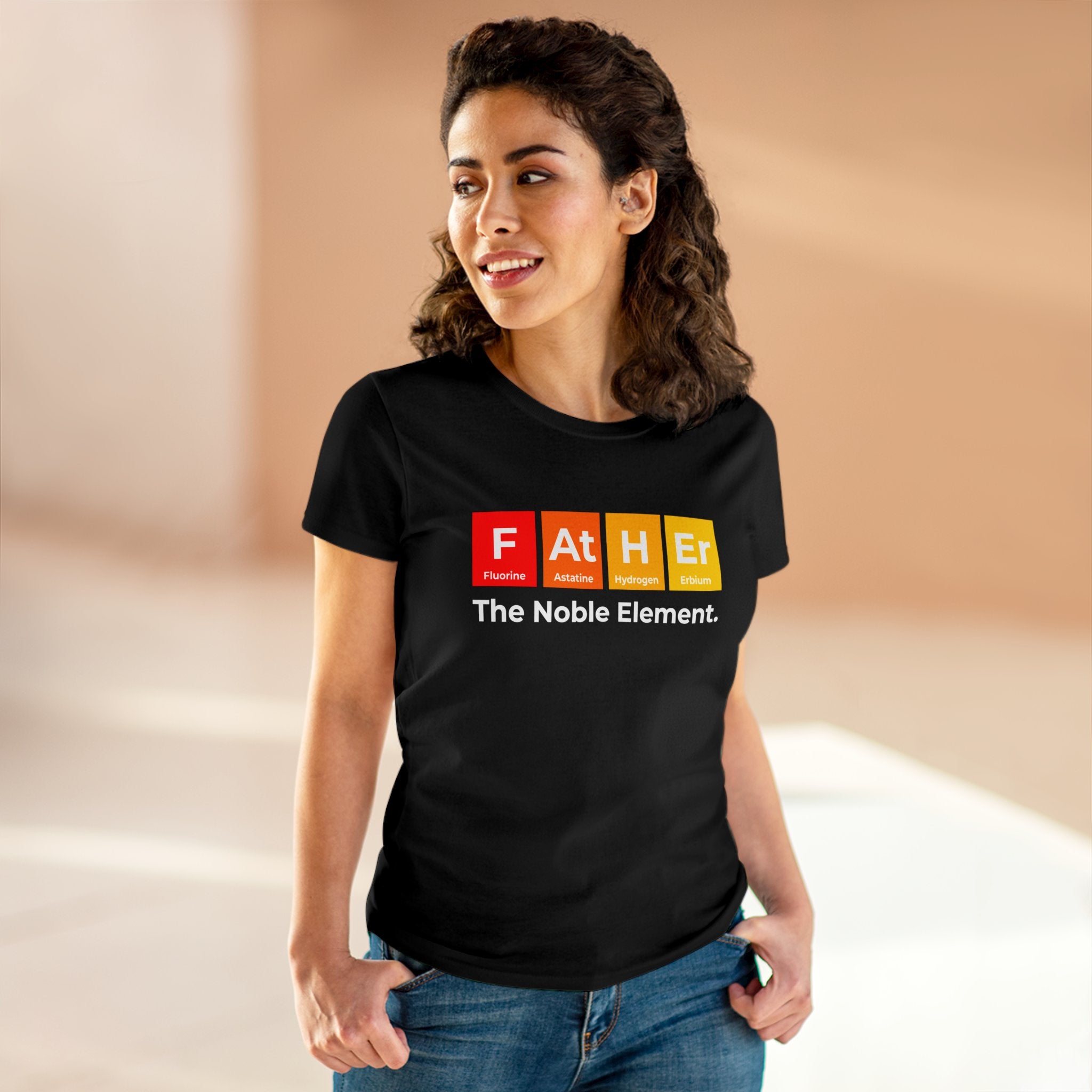 Woman wearing a black, 100% cotton t-shirt with a design mimicking periodic table elements spelling "Father" and the phrase "The Noble Element." She is standing indoors with a neutral background, showcasing the Father Graphic - Women's Tee that offers warm and cozy comfort.