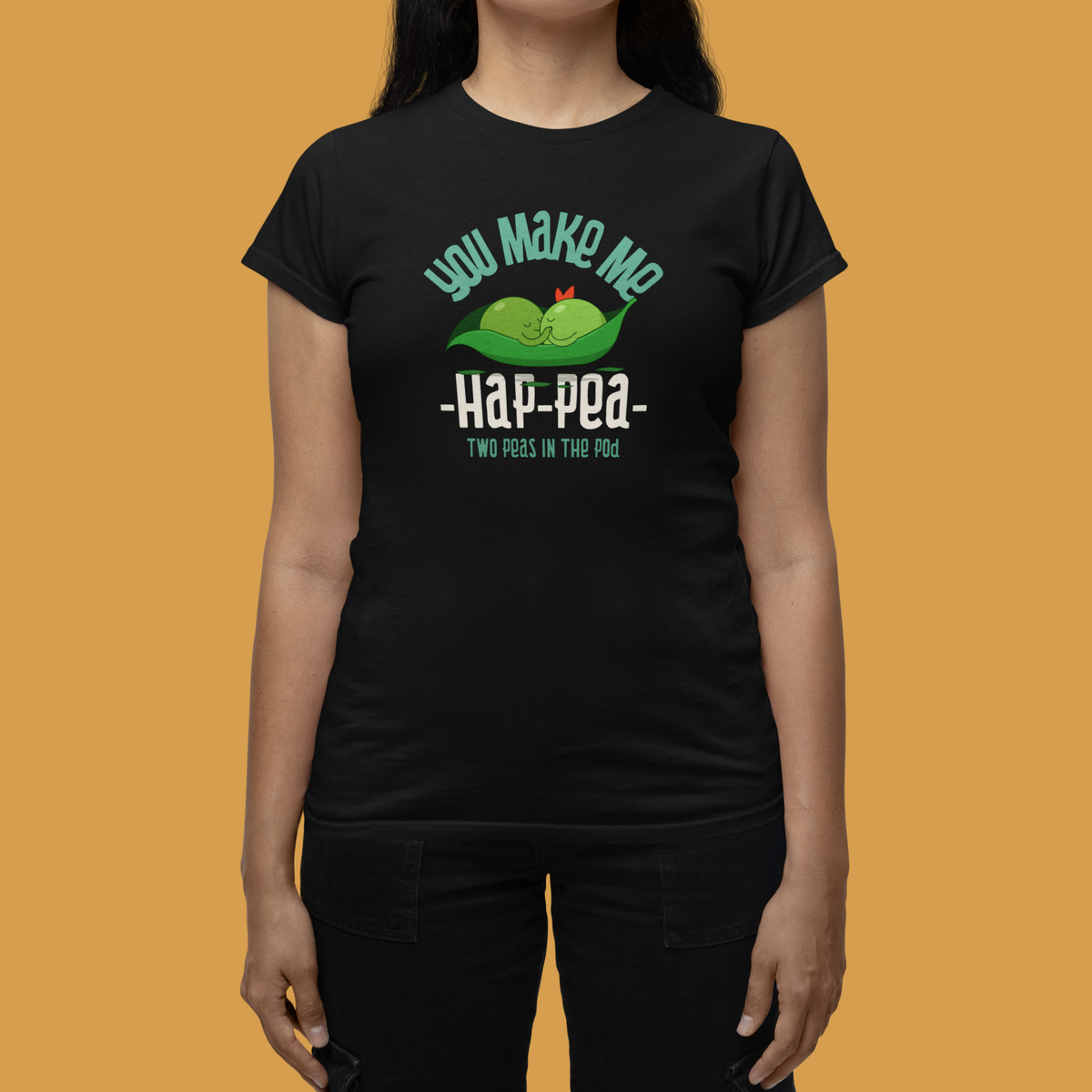 'You Make Me Hap-Pea' t-shirt in black, close-up of woman wearing it against orange backdrop, cute pea pun graphic tee