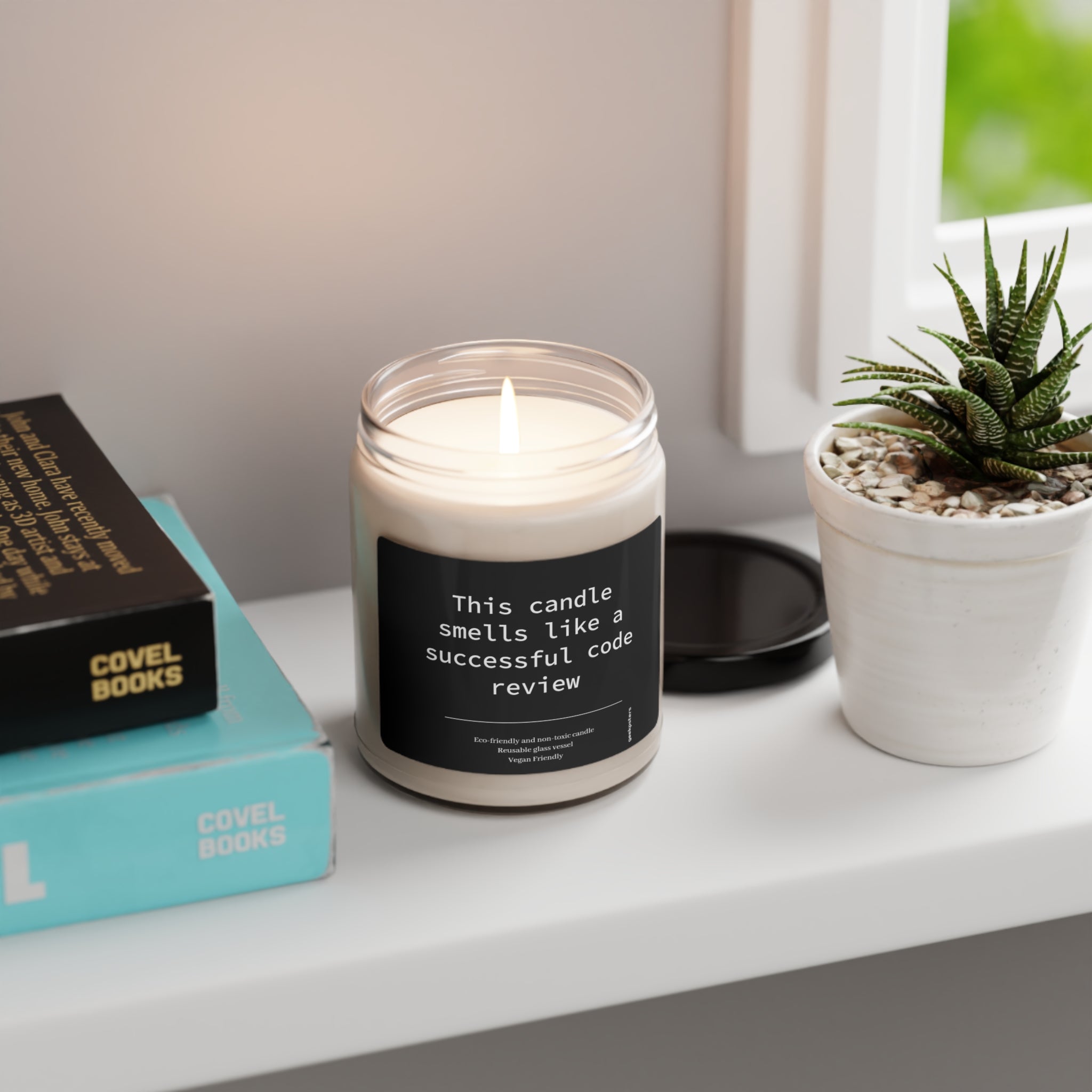 A lit This Candle Smells like a Successful Code Review - Scented Soy Candle, 9oz labeled "this candle smells like a successful coding session" on a windowsill, next to books and a potted succulent.
