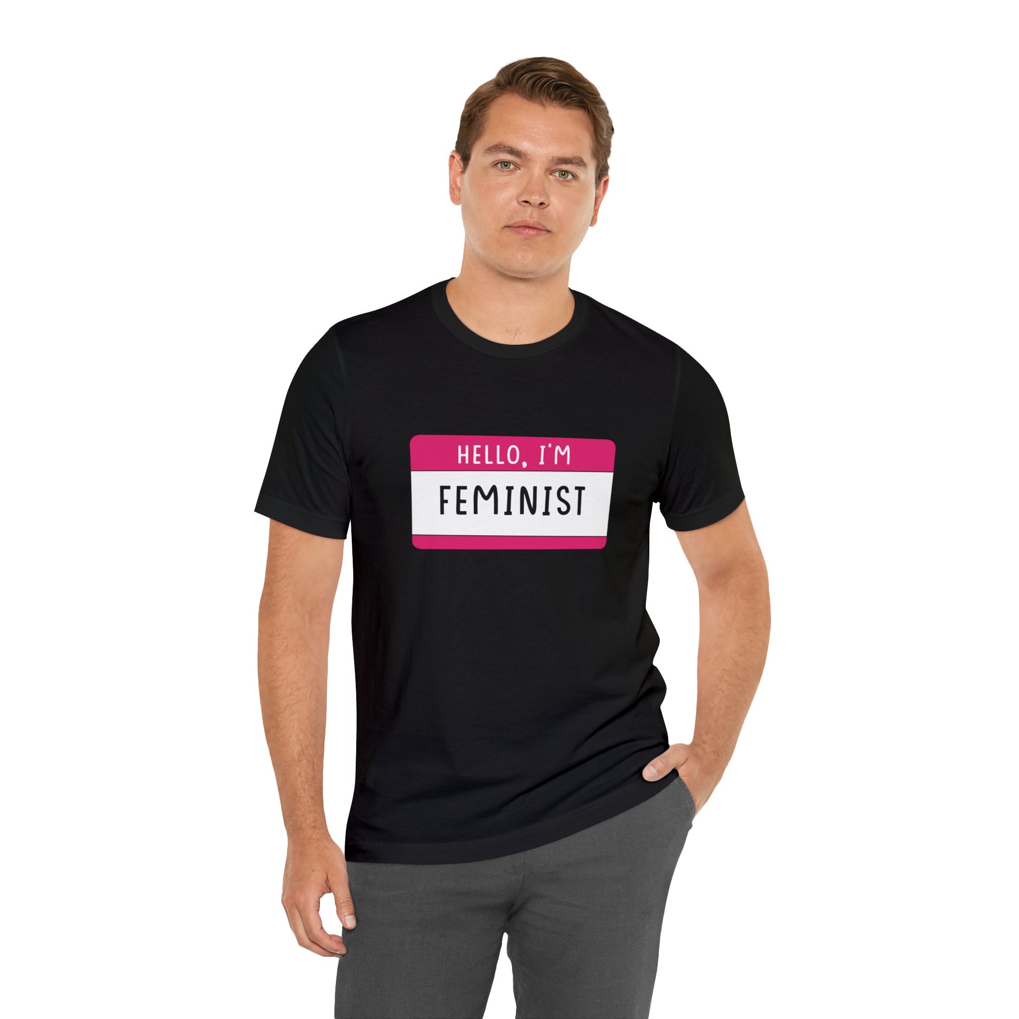 Man in a black Hello, I'm Feminist T-Shirt with "hello, i'm feminist" printed in white and pink on the front, standing against a plain background.