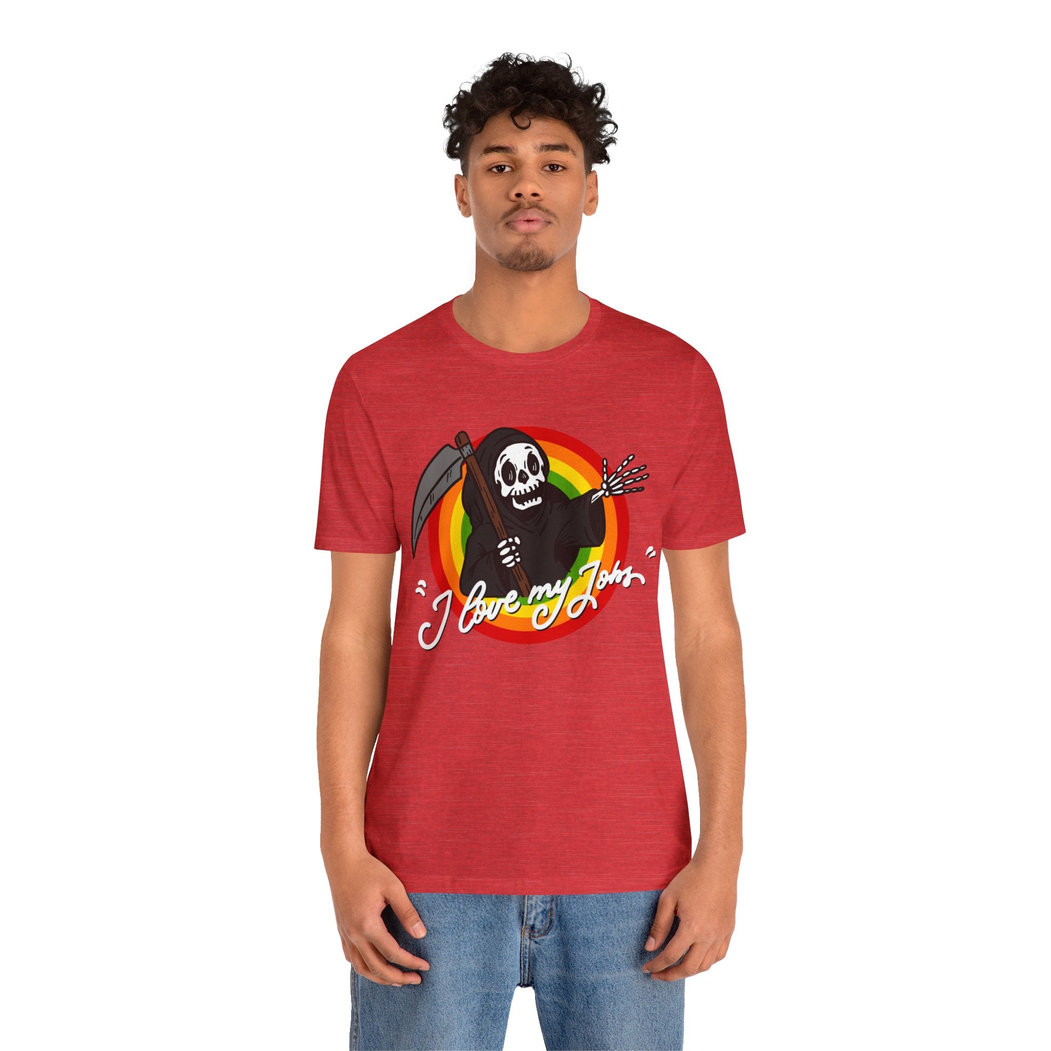 Sentence with product name: A young man wearing a red Love My Jobs T-Shirt with a skull, crossbones, and "Love My Job" graphic, paired with blue jeans, standing against a plain background.