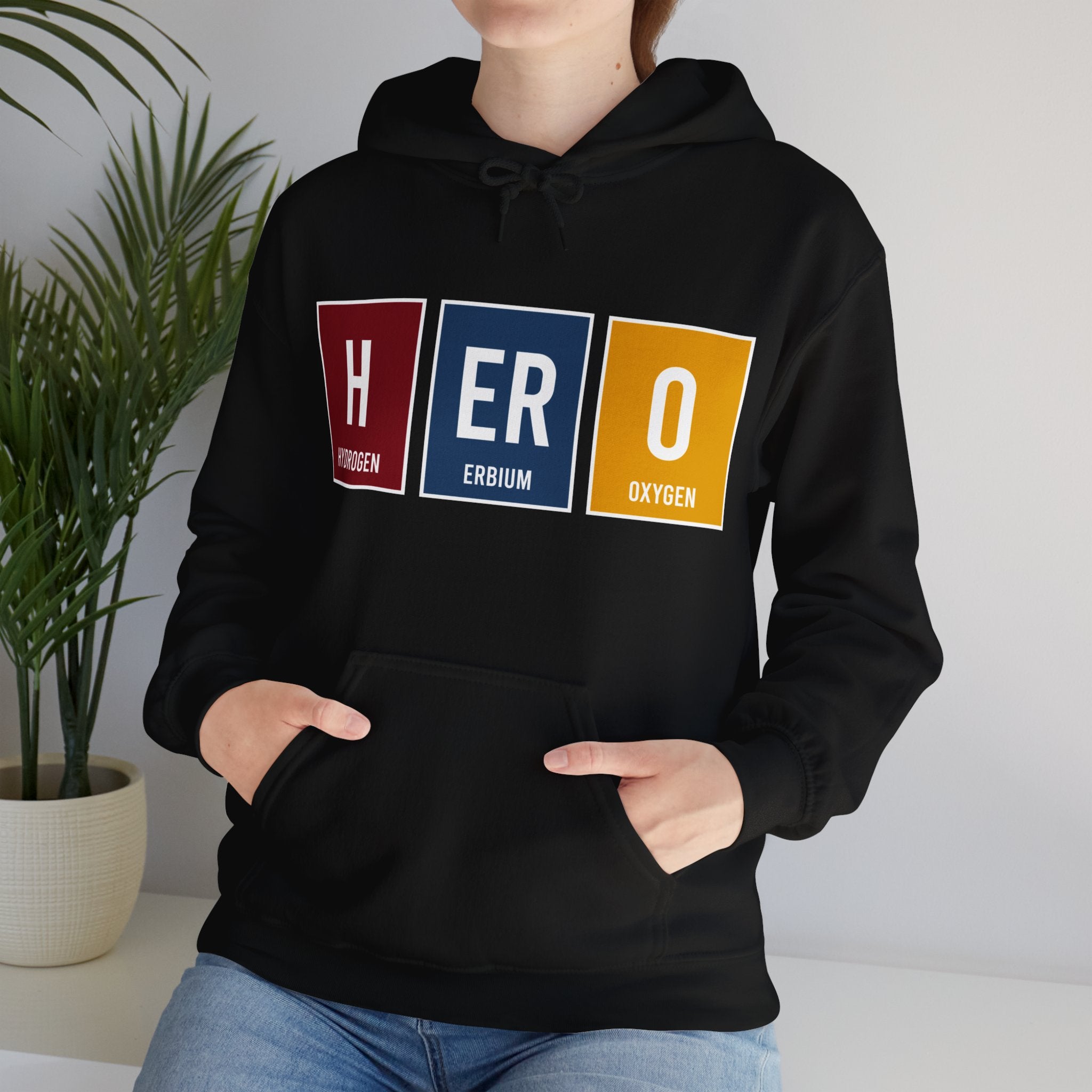 Person wearing a black HERO - Hooded Sweatshirt with the word "HERO" displayed using periodic table element symbols for Hydrogen, Erbium, and Oxygen. Hands are in the front pocket. Plant in the background underscores their quiet heroism.