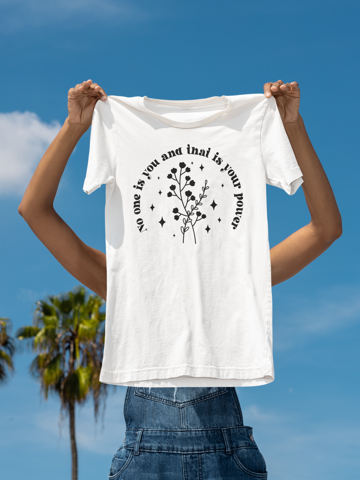 'No One Is You And That Is Your Power' t-shirt in white, held up on a beach