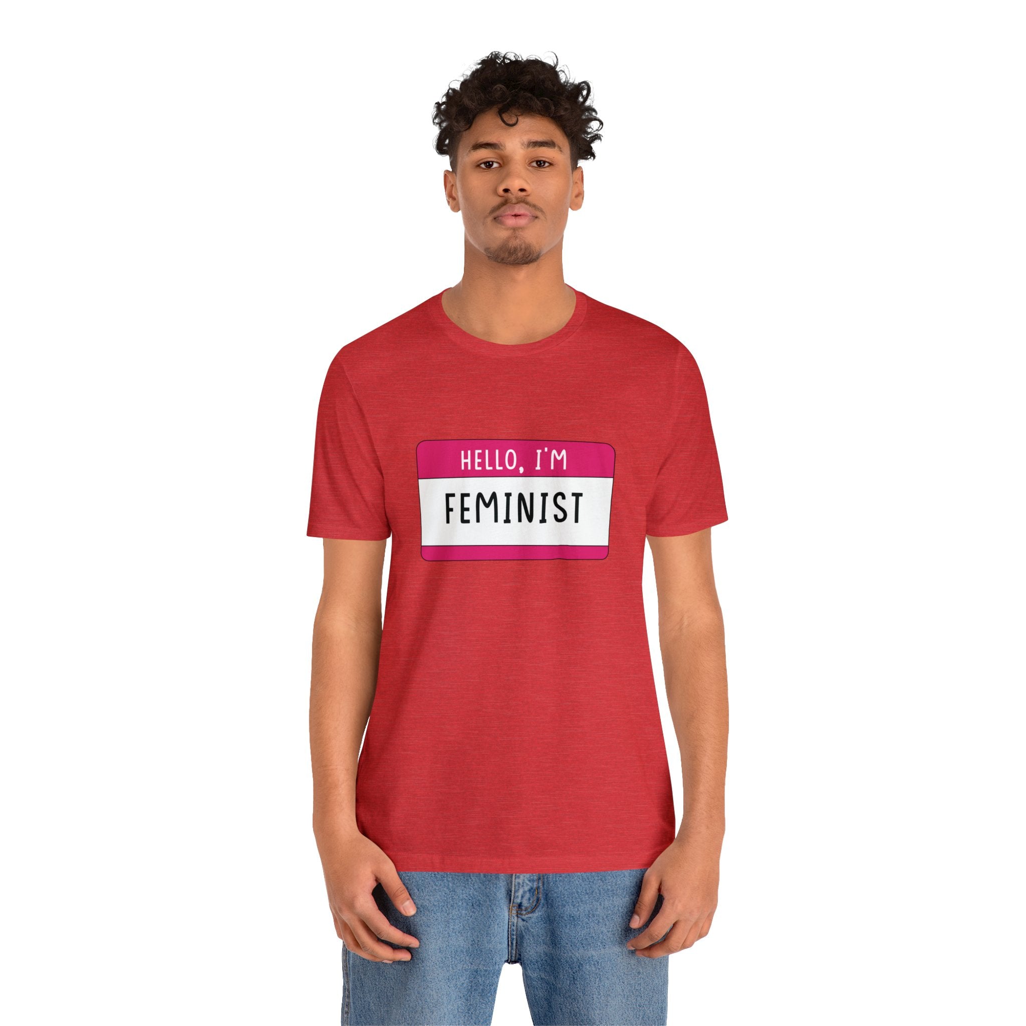 A young man in a red Hello, I'm Feminist T-Shirt stands facing the camera.
