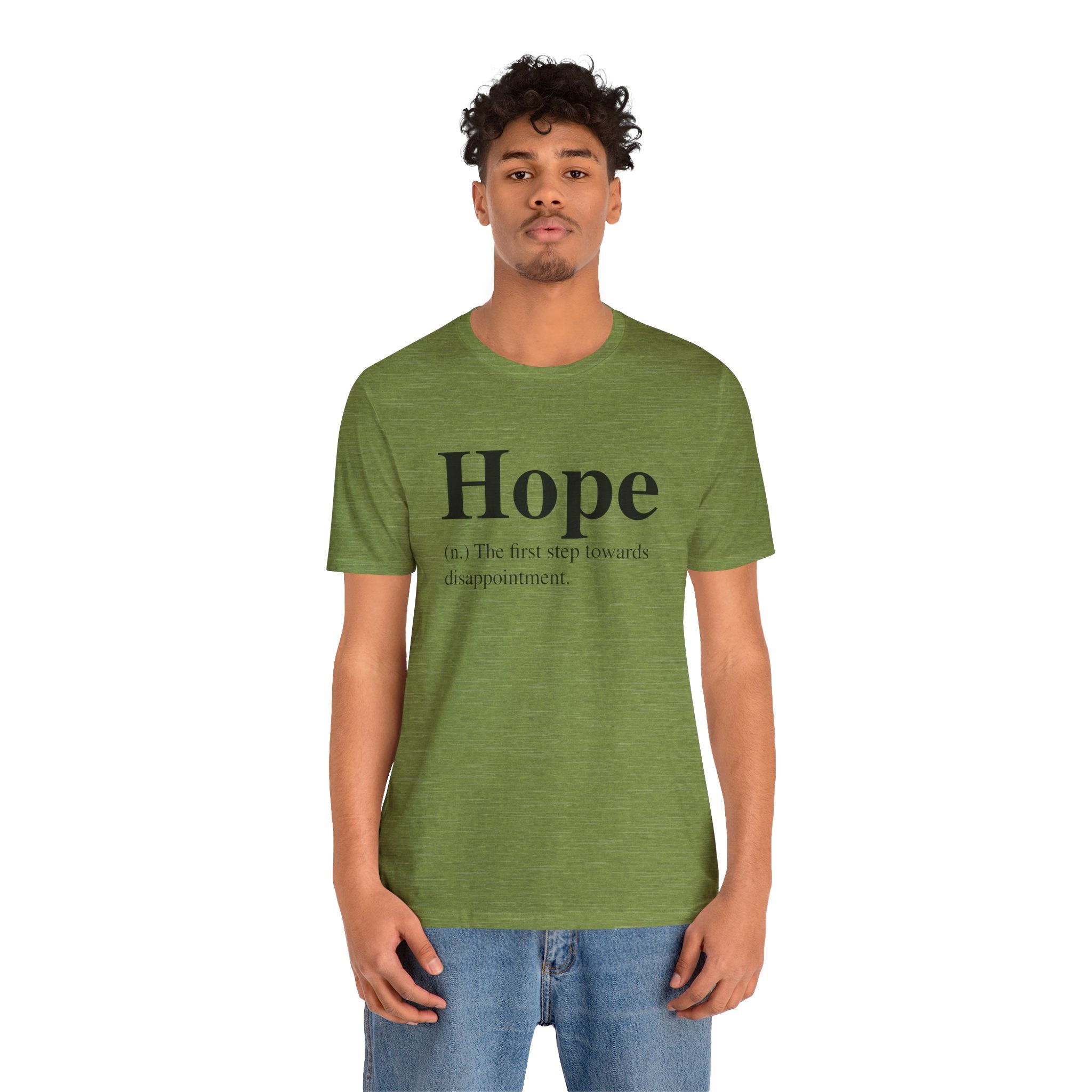 Man in a green unisex "Hope T-Shirt" with the word "hope" and a cynical definition printed on it, paired with blue jeans, standing isolated against a white background.