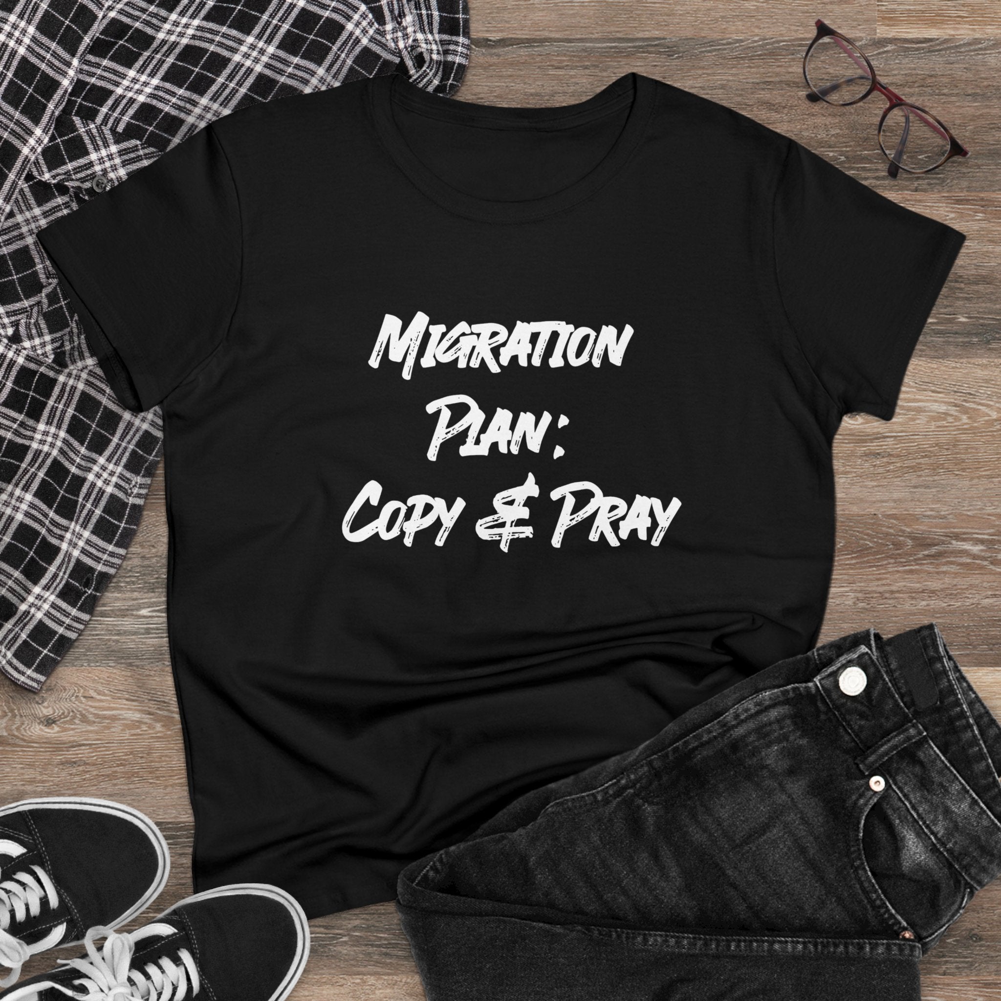 The "Migration Plan Copy and Pray" Women's Tee, crafted from black cotton and featuring a unique design, is stylishly displayed on a wooden floor with a plaid shirt, glasses, and sneakers as accents.