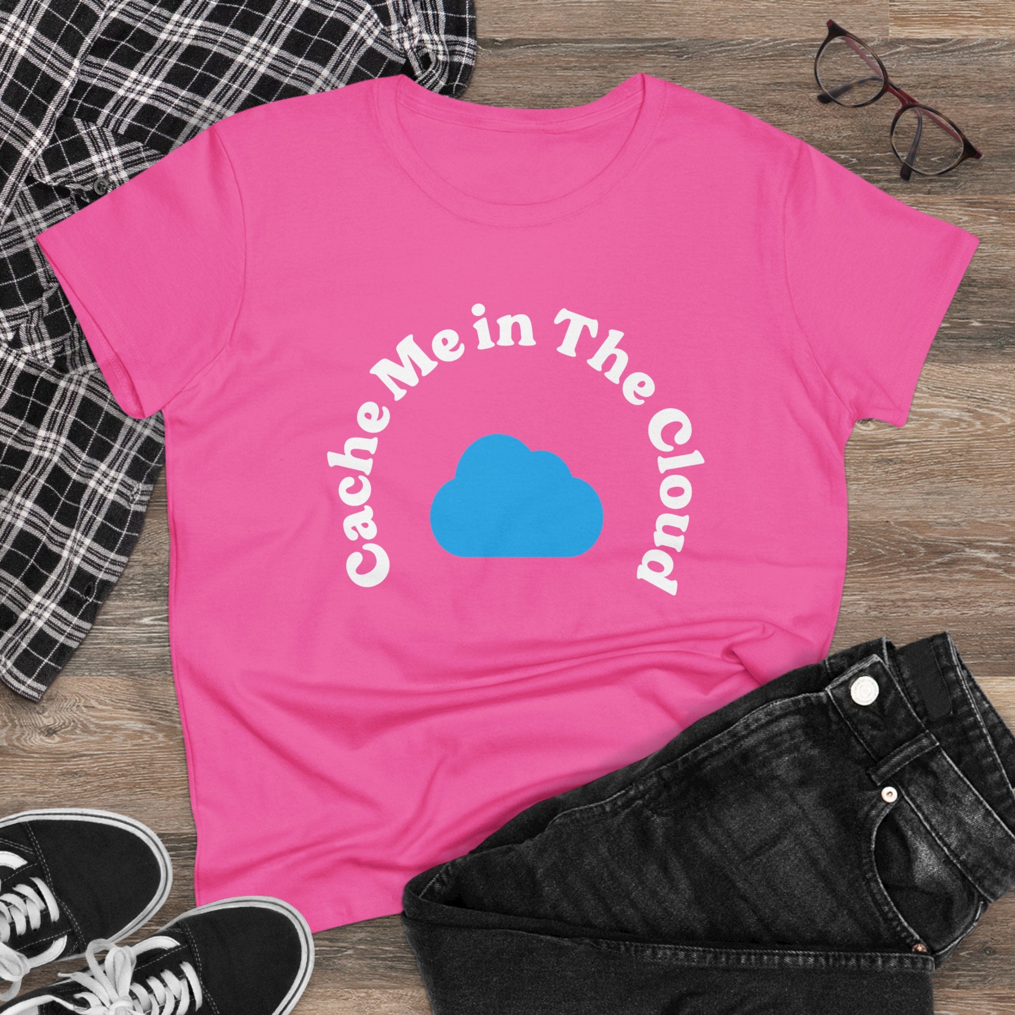 The "Cache Me in The Cloud" Women's Tee is crafted from light cotton and showcases a playful blue cloud graphic with text. It's styled on a wooden surface alongside black jeans, a plaid shirt, glasses, and sneakers for a chic look.