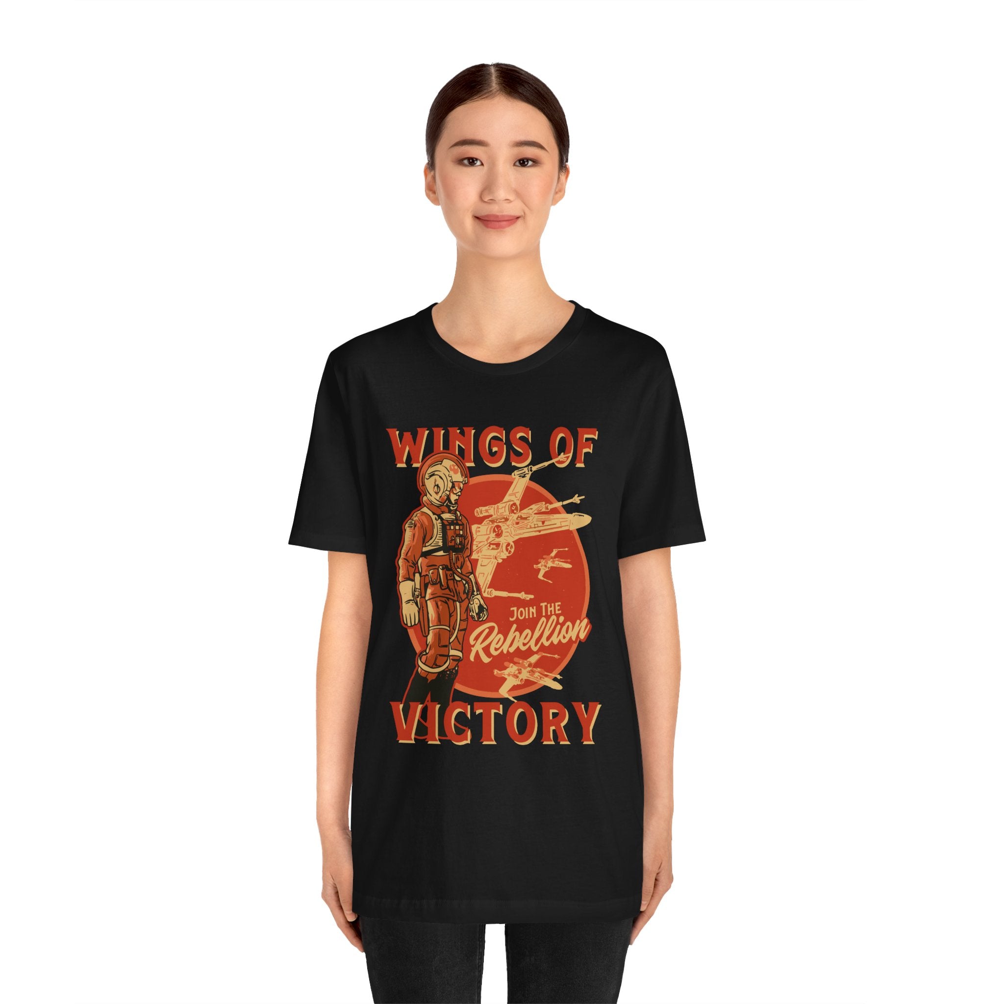 A young woman wearing a black unisex Wings of Victory T-shirt with a vintage orange and yellow graphic that reads "wings of rebellion victory.