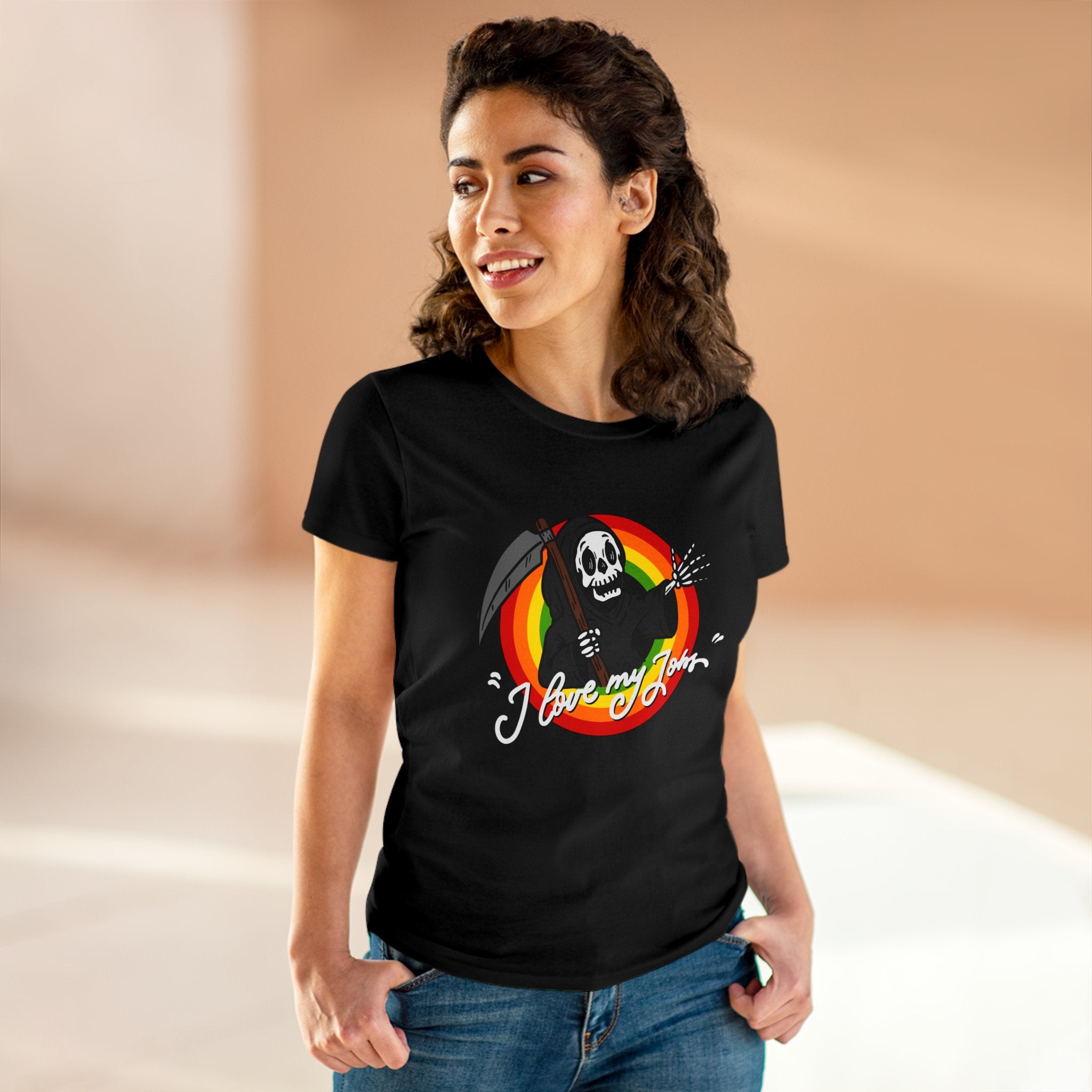 A woman with wavy hair stands indoors wearing the soft, comfortable cotton Love My Jobs - Women's Tee featuring a scythe-wielding skeleton graphic and the text "I love my job." She has her hands in her pockets and is smiling.