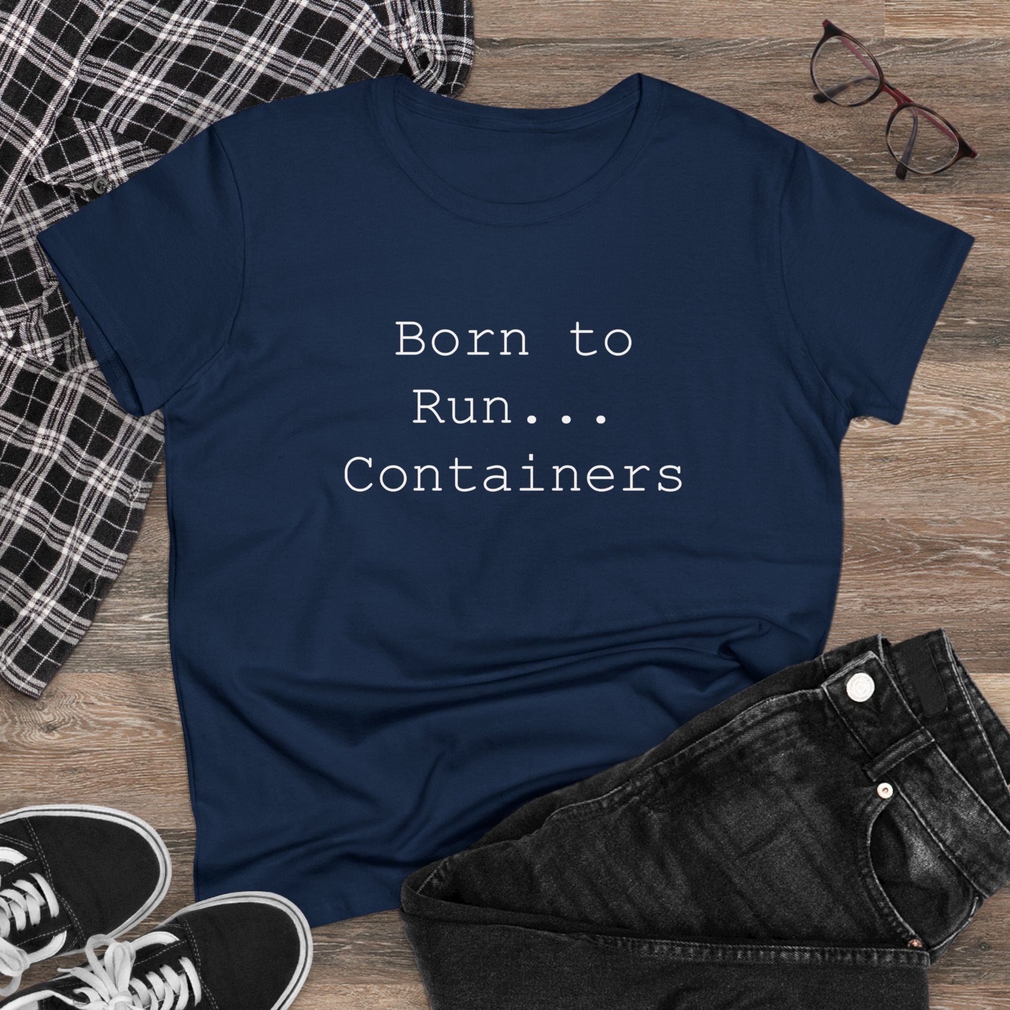 The "Born to Run Containers - Women's Tee," in navy blue, is displayed on a wooden floor alongside a plaid shirt, glasses, and black sneakers. This wardrobe staple beautifully marries comfort with style.