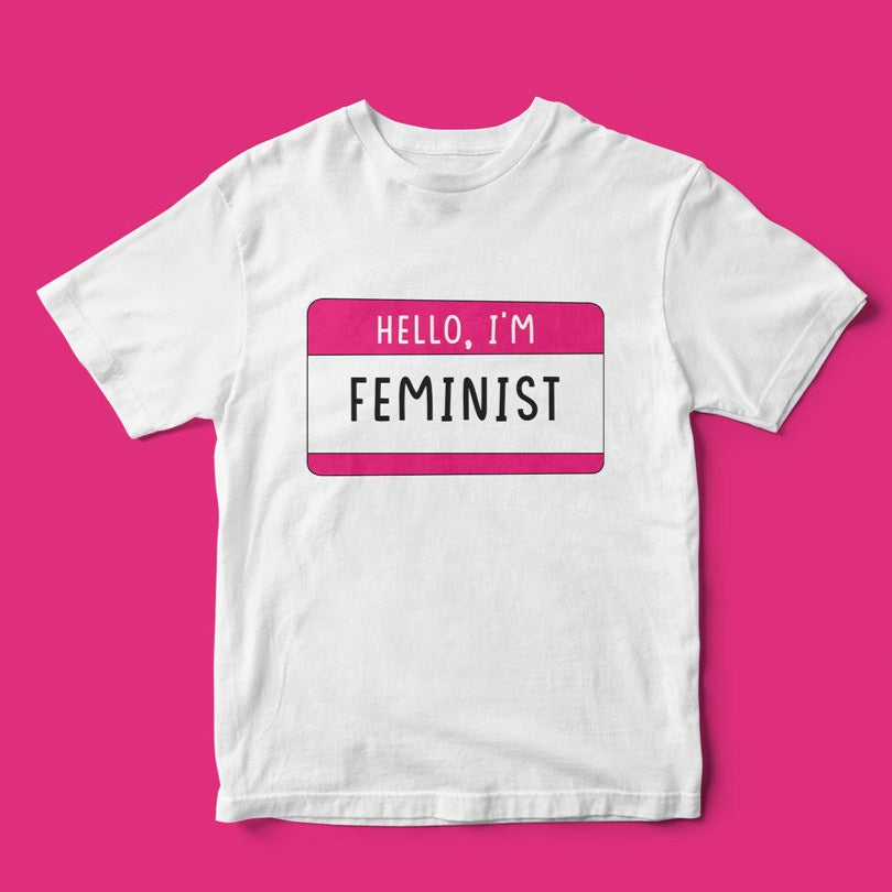 Hello, I'm Feminist T-Shirt with a "hello, I'm feminist" name tag design celebrating equality, laid flat on a pink background.