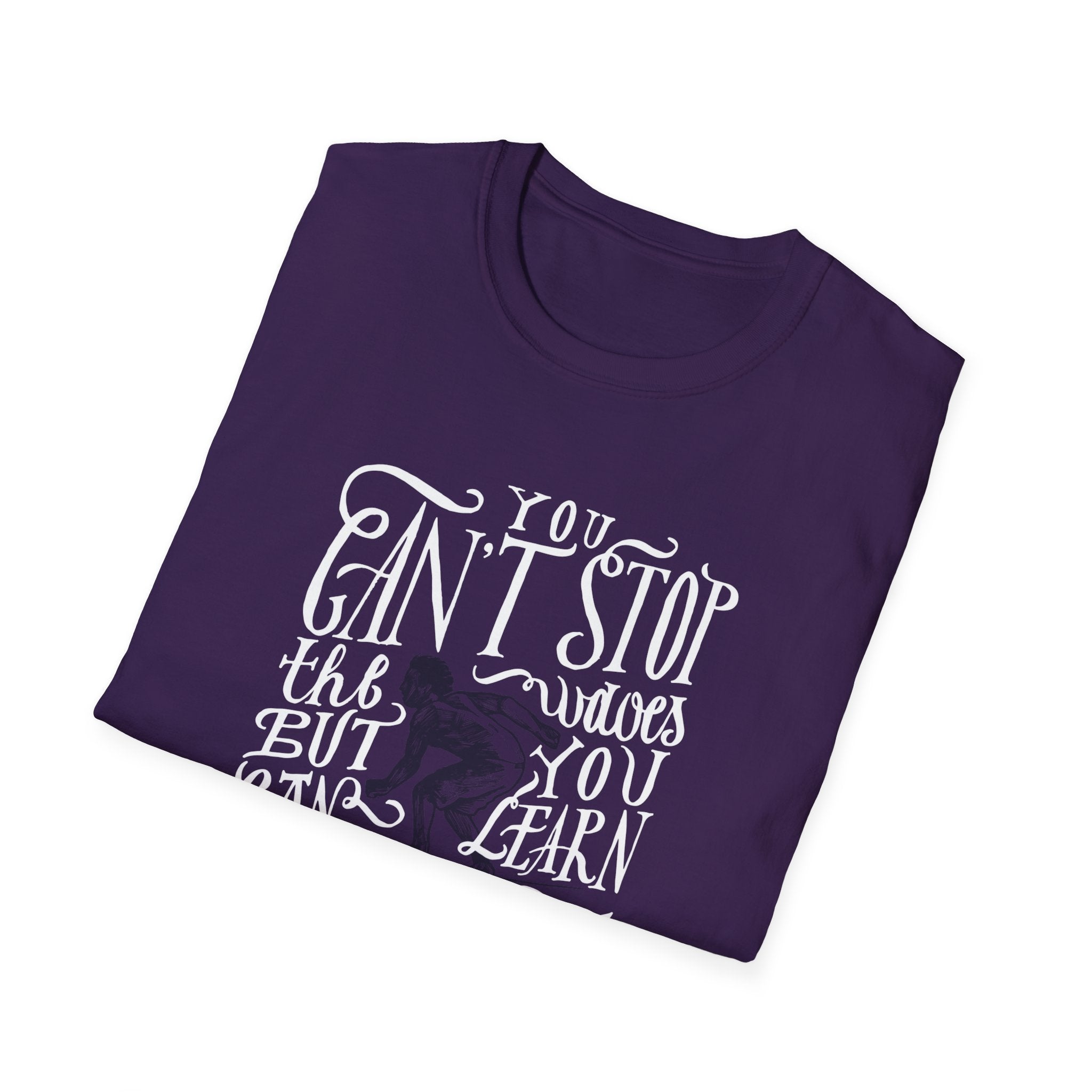 The folded Can't Stop Surfing T-Shirt, showcasing a vintage-inspired design, features white lettering with the phrase "You can't stop the waves, but you can learn to surf." Ideal for those who embrace resilience and style, this purple surfing t-shirt is a must-have wardrobe essential.