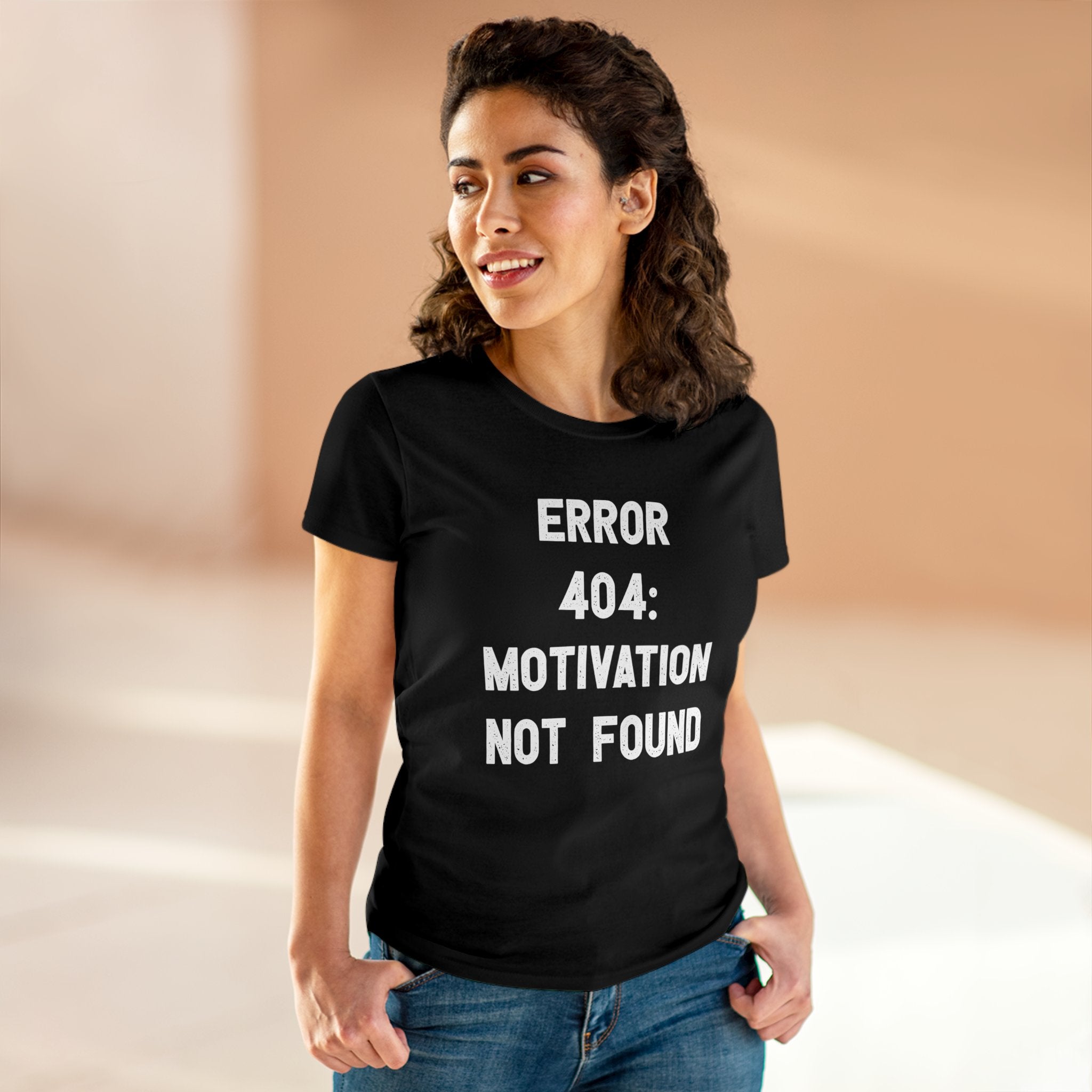 A woman is standing indoors, wearing a black "Error 404: Motivation not found - Women's Tee" that reads "ERROR 404: MOTIVATION NOT FOUND" in white text. Made of pre-shrunk cotton, it features a semi-fitted silhouette. She is smiling and looking to her right.


