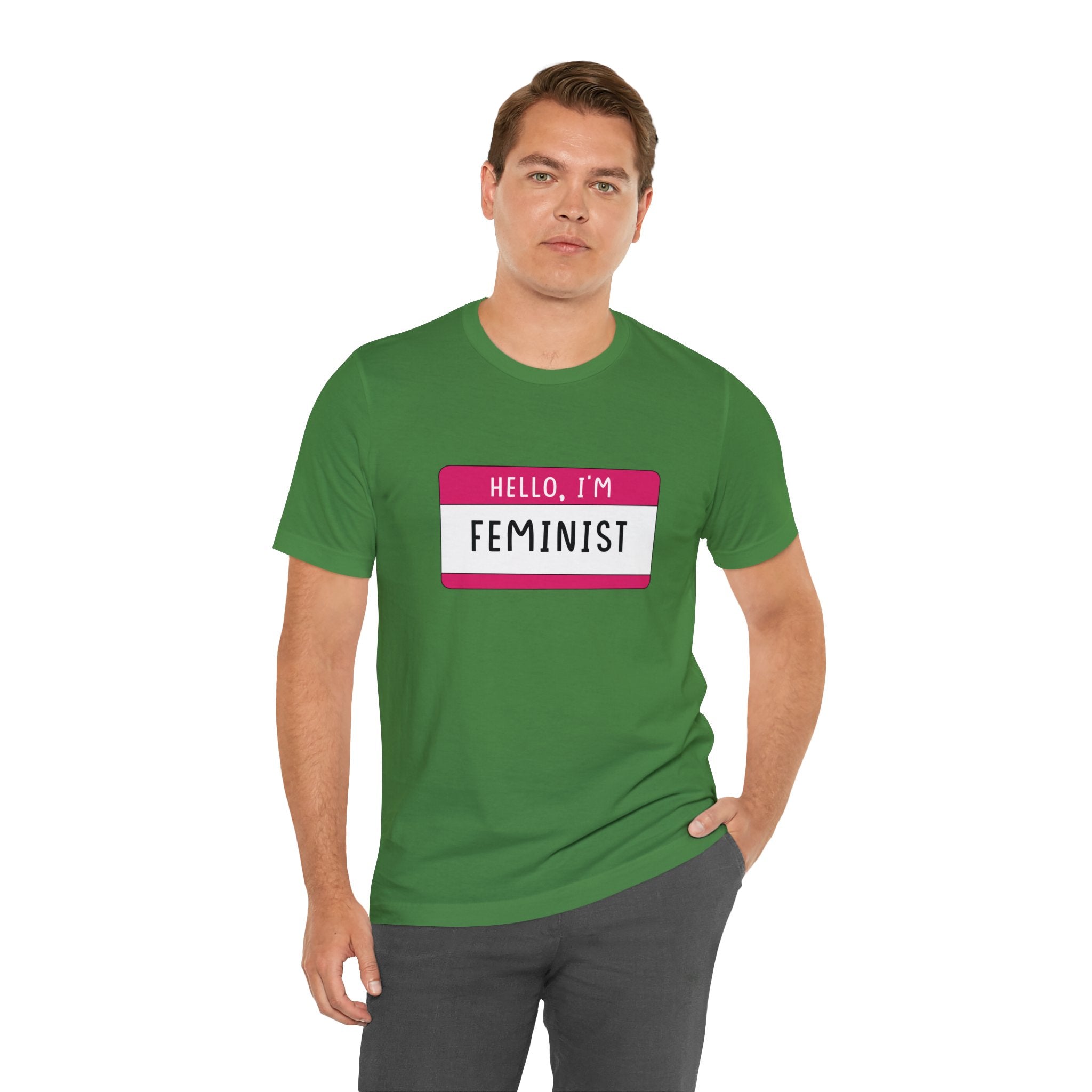 Man in a green Hello, I'm Feminist T-Shirt with a "hello, i'm feminist" badge design, standing against a white background.