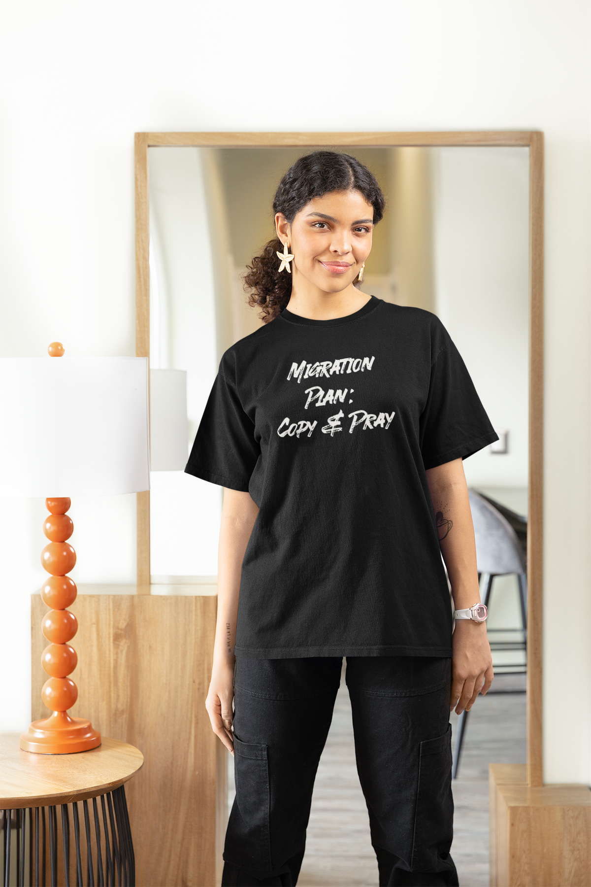 'Migration Plan: Copy and Pray' women's tee in black, woman standing indoors at home, funny developer gift shirt