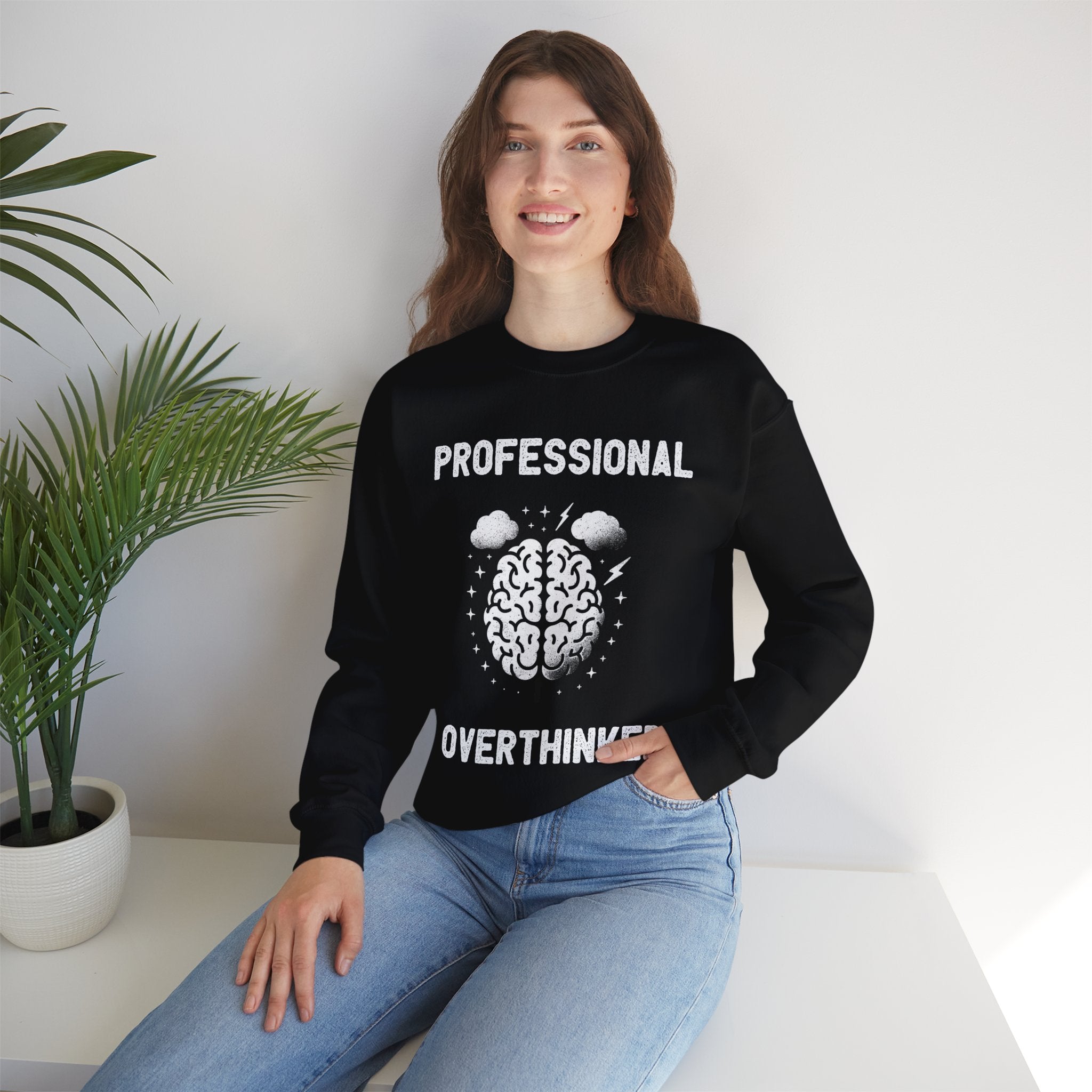 A woman smiling, wearing a Professional Overthinker - Sweatshirt that reads "Professional Overthinker" with an illustration of a brain. She is seated next to a potted plant, looking comfy and cool in the perfect warmth for colder months.