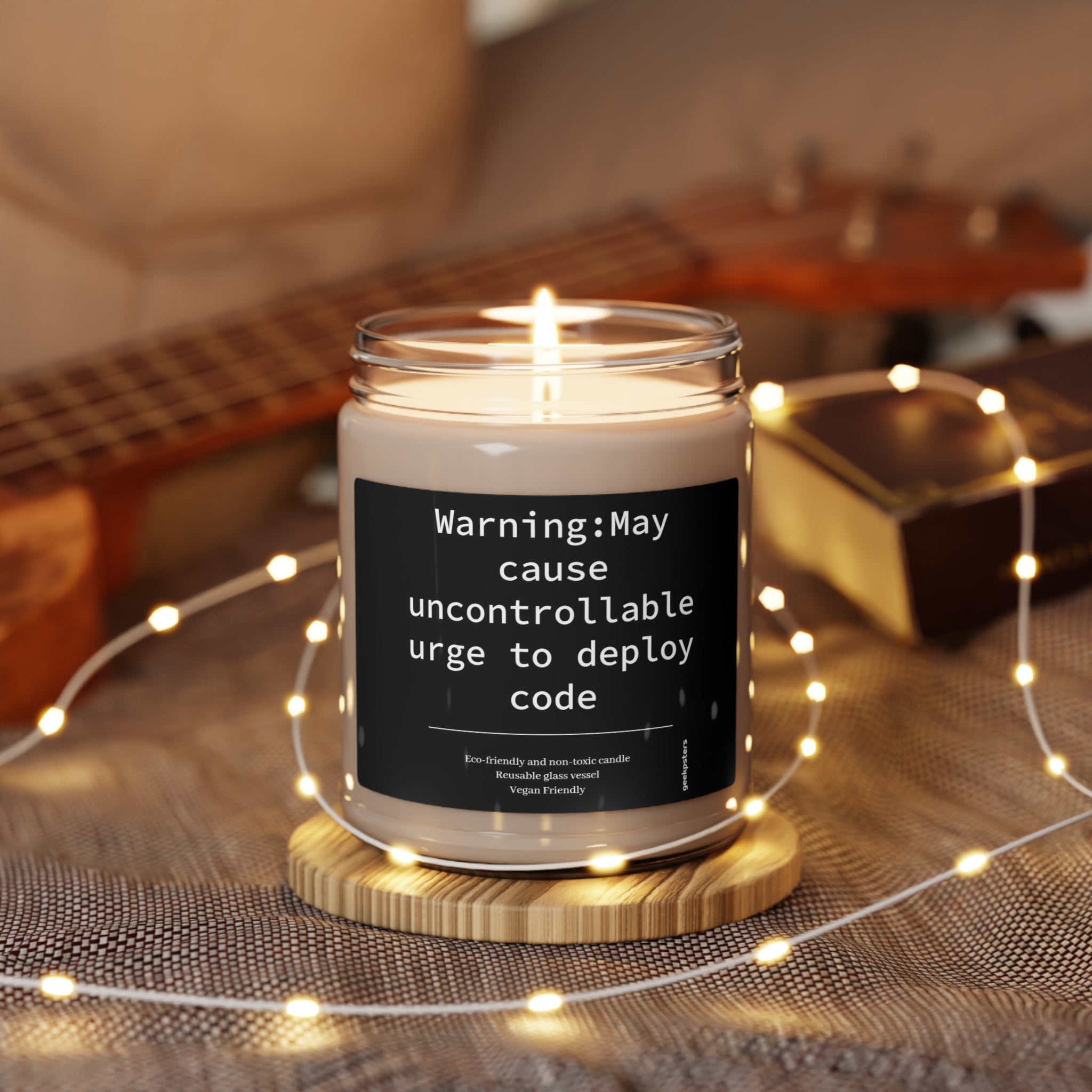 A Caution May Cause Urge to Deploy - Scented Soy Candle, 9oz with a humorous label warning that it may cause an uncontrollable urge to deploy code, placed on a table surrounded by string lights.