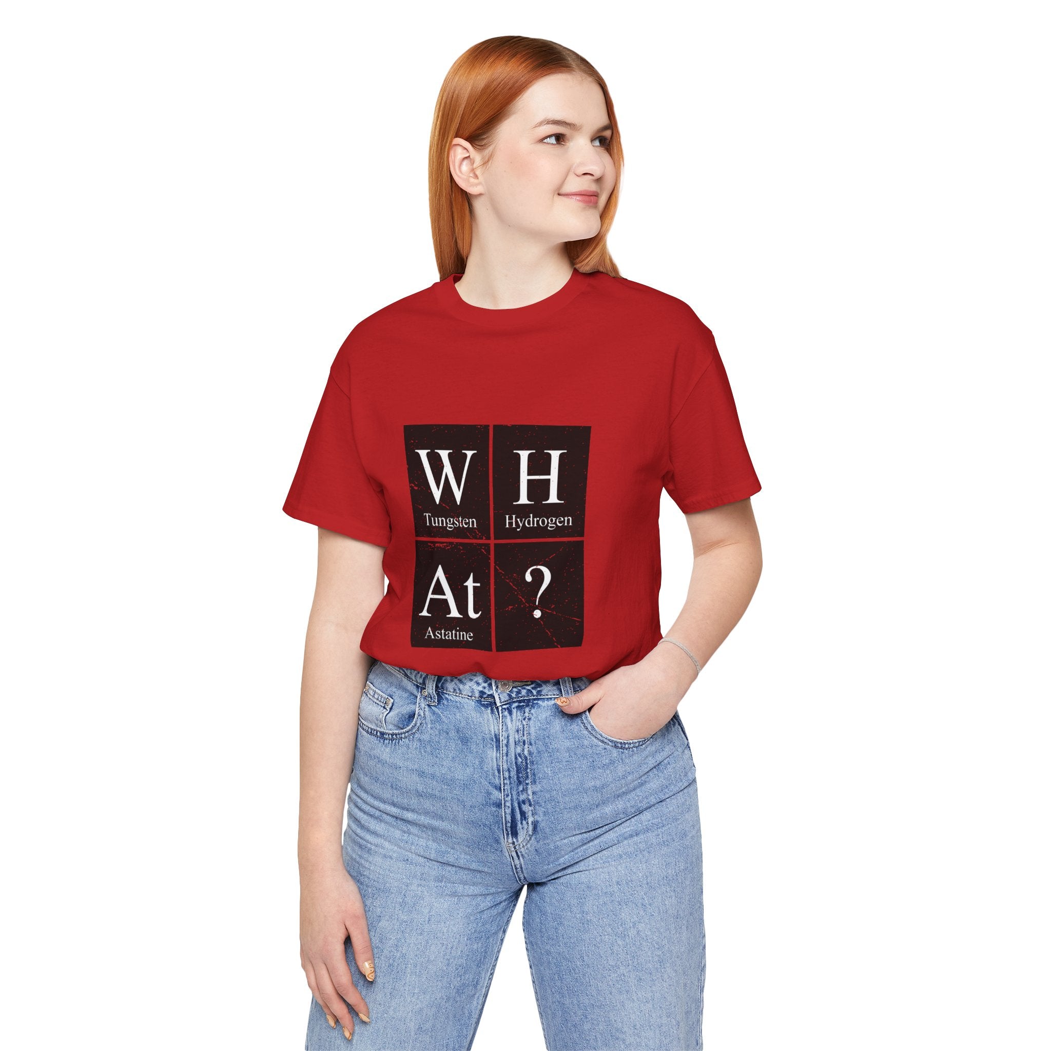 A woman in a red unisex jersey tee with "W-H-At-?" printed on it, pairing it with light blue jeans, stands with hands in pockets, looking to her right.