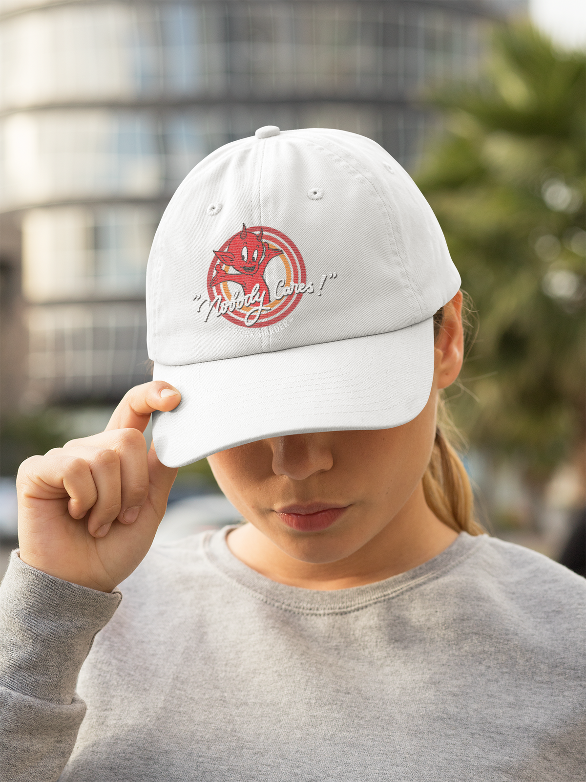 "Nobody Cares!" red devil graphic dad hat in white, woman wearing it outdoors, funny sarcastic gift cap