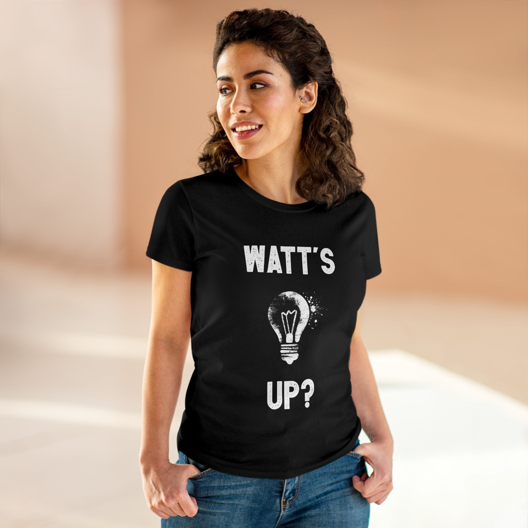 A woman wearing a black t-shirt that reads "Watt's Up?", featuring a light bulb graphic, is standing in a well-lit indoor space. She is smiling and has her hands in her pockets, enjoying the comfort of the light cotton fabric Watts Up - Women's Tee, which is also pre-shrunk for a perfect fit.