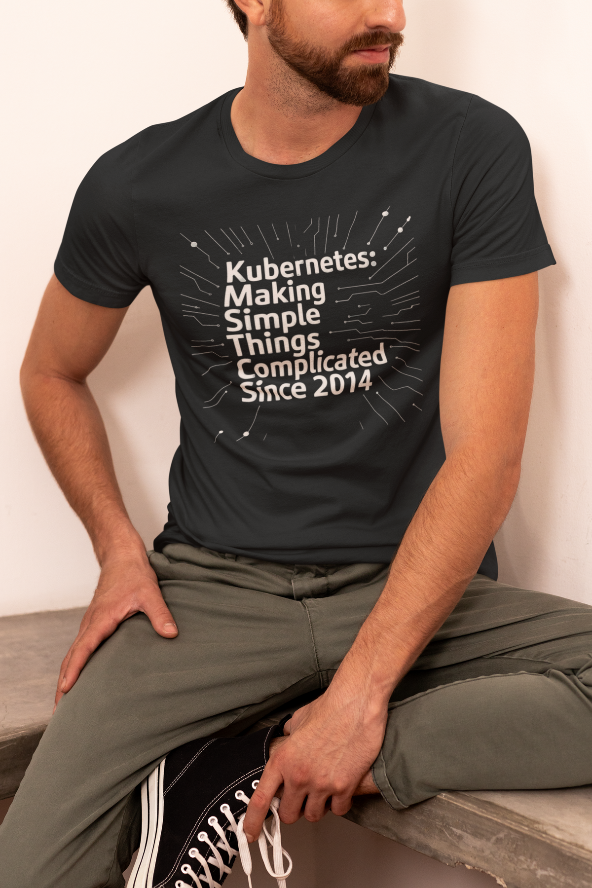 'Kubernetes: Making Simple Things Complicated Since 2014' t-shirt in black, man wearing it seated, funny DevOps gift