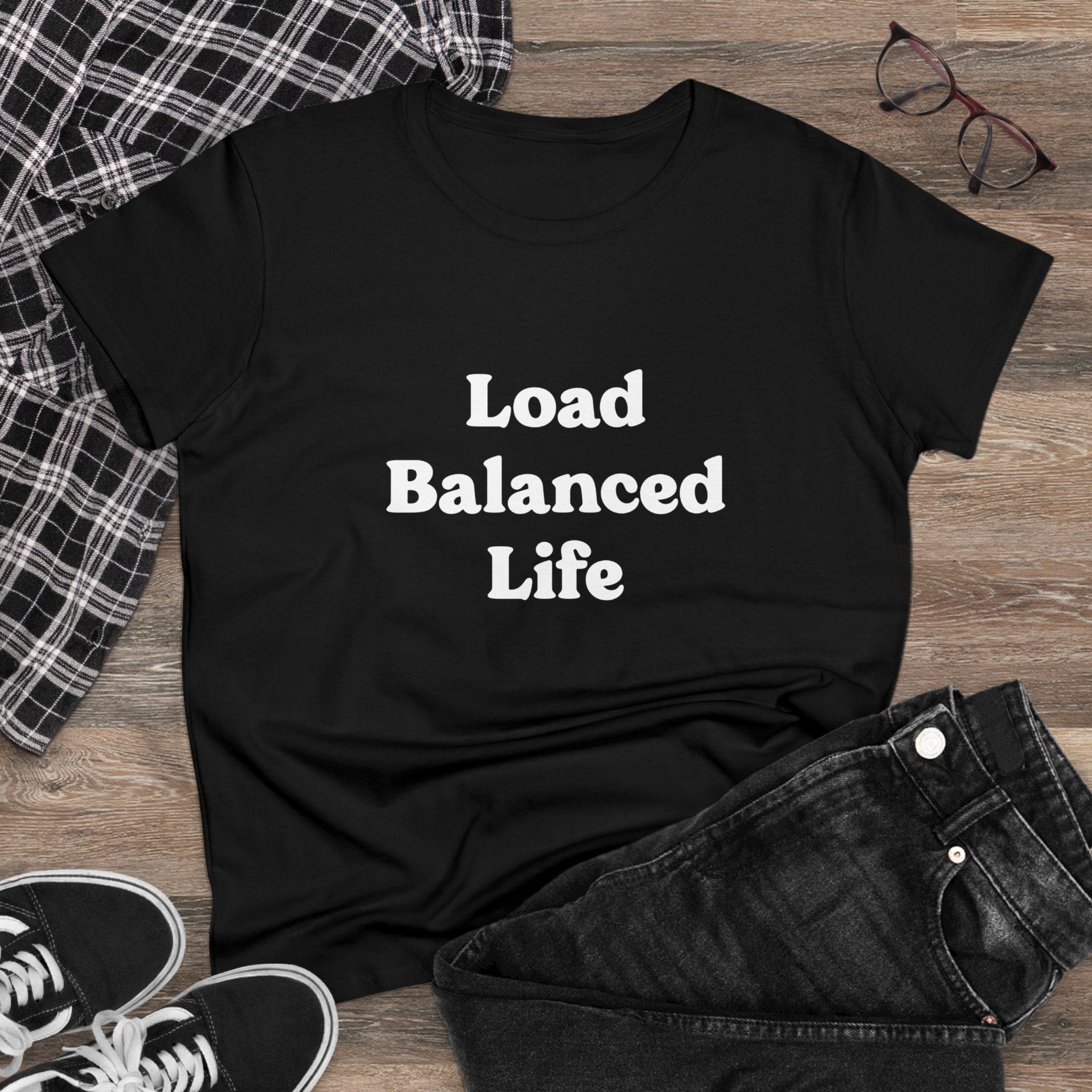 The stylish and comfortable Load Balanced Life - Women's Tee, featuring "Load Balanced Life" text, lies on a wooden floor. It's placed among a plaid shirt, black jeans, glasses, and sneakers. Made from pre-shrunk cotton, this black t-shirt ensures the perfect fit every time.