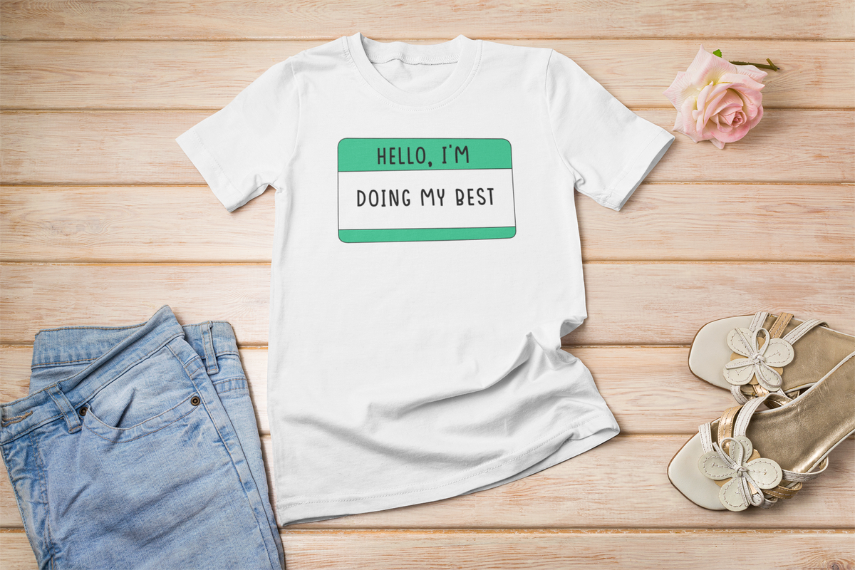 Hello I'm Doing My Best name tag T-shirt in white, flat lay styled with roses on wood, positive gift tee