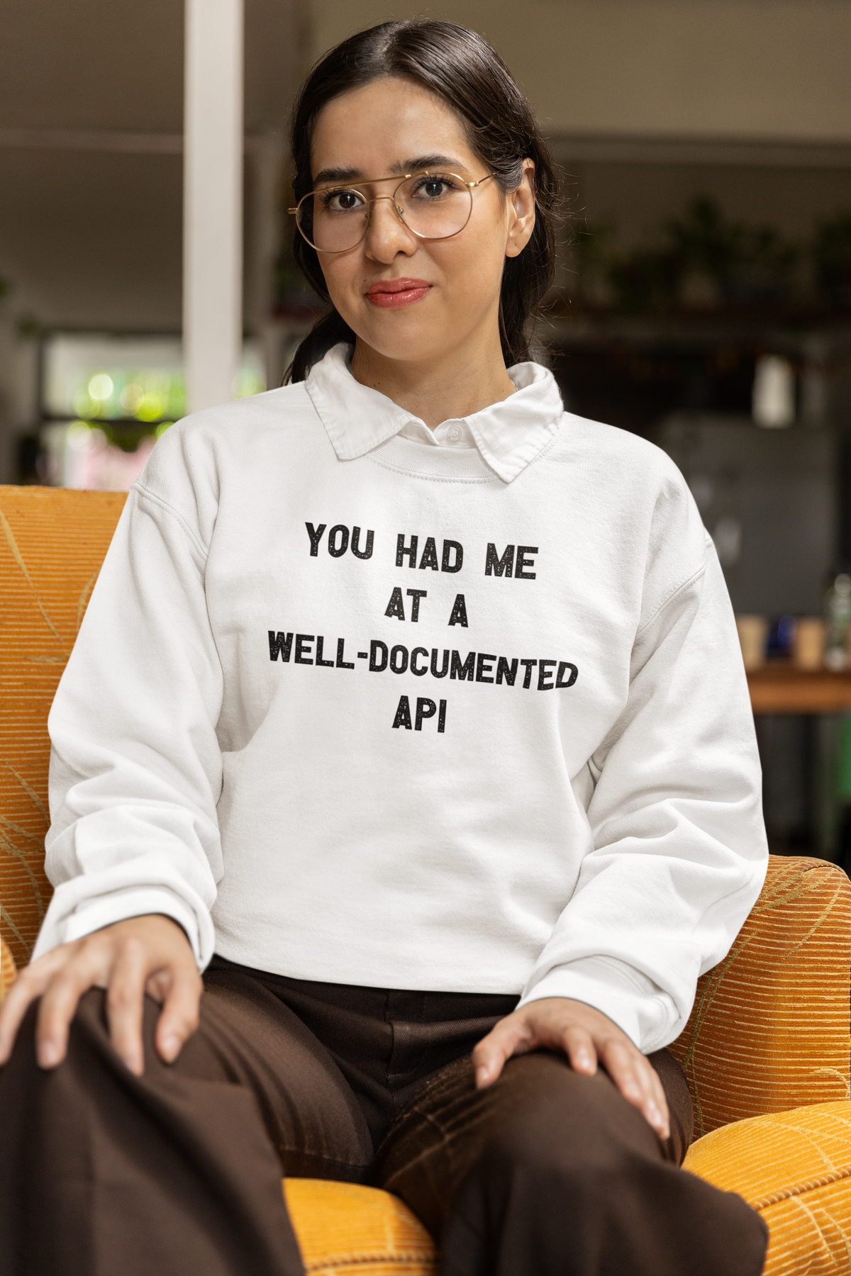 "You Had Me At A Well-Documented API" sweatshirt in white, woman wearing it seated in glasses, funny coder gift