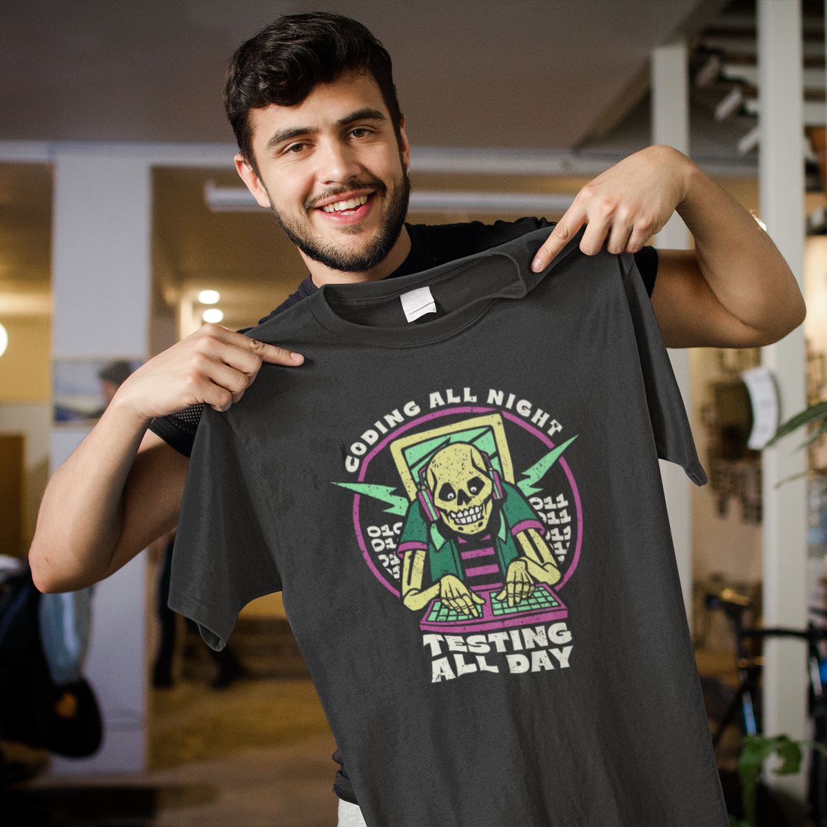'Coding All Night, Testing All Day' skeleton t-shirt in black, man model holding it up indoors, funny gift for coders