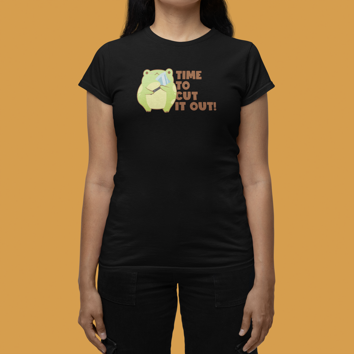 'Time to Cut It Out' frog women's tee in black, flat lay on mustard backdrop, funny pun gift idea