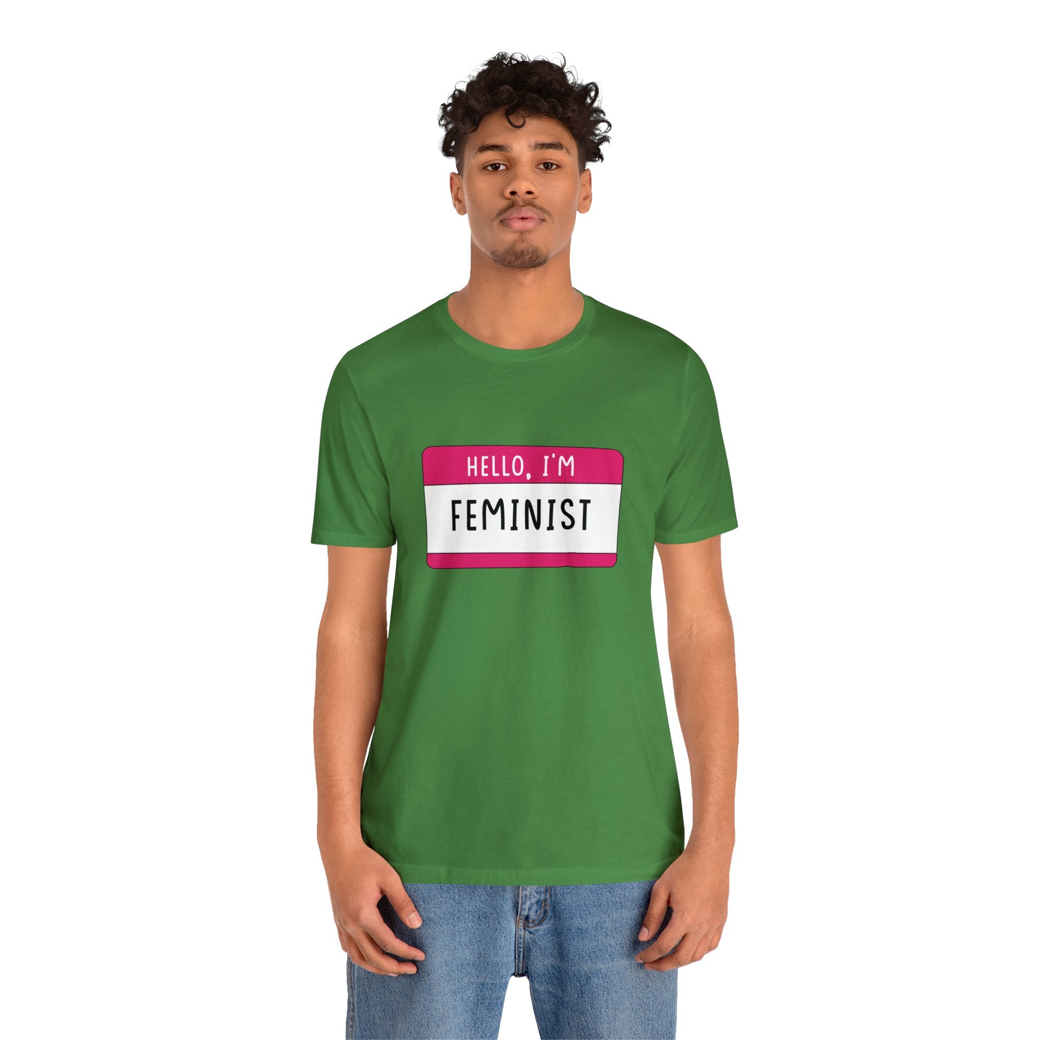 Young man wearing a green Hello, I'm Feminist T-Shirt with a "hello, I'm feminist" badge design, standing against a white background.