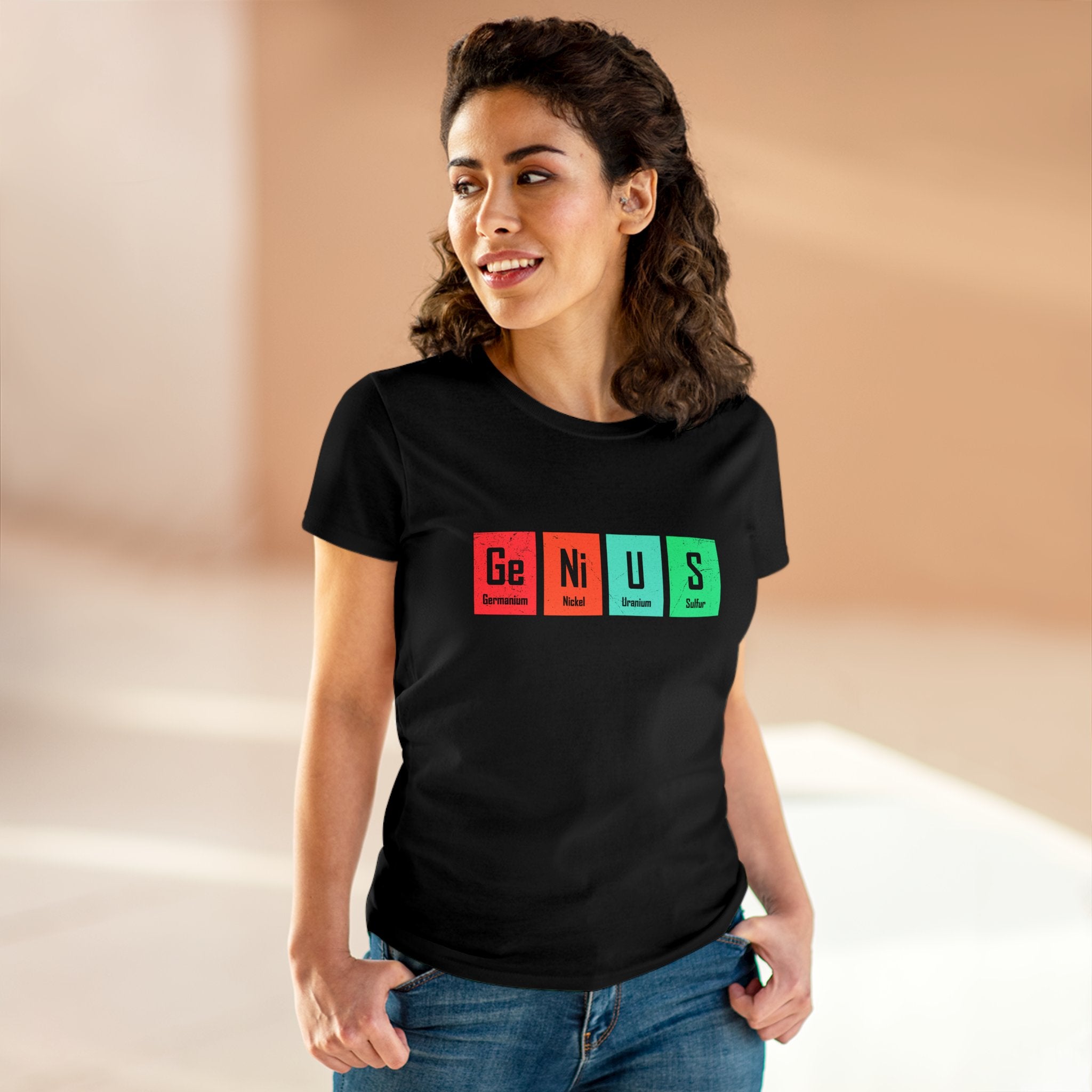 A woman wearing a cozy and stylish black Ge-Ni-U-S - Women's Tee with the design spelled out using periodic table elements stands indoors, casually smiling with her hands in pockets.