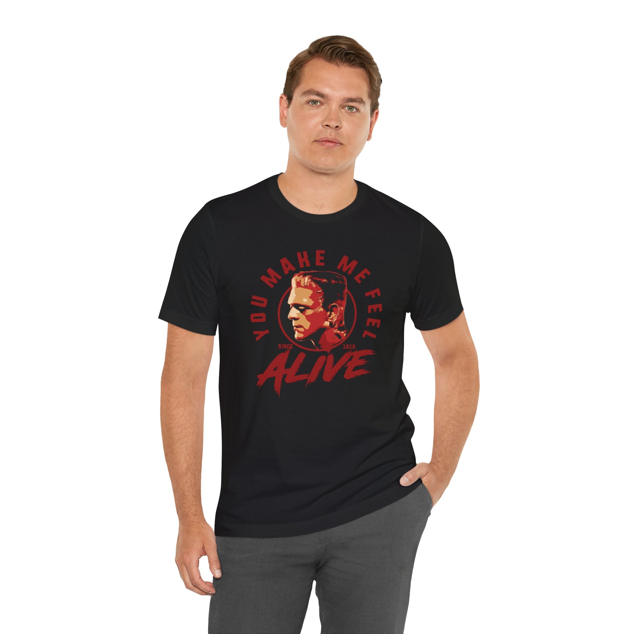 Man wearing a black You Make Me Feel Alive unisex tee with red and yellow quality print, standing against a plain background.