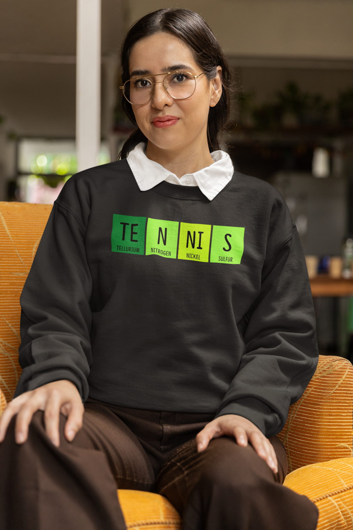 'Tennis' periodic table sweatshirt in black, woman wearing it seated indoors, funny chemistry gift for tennis fans