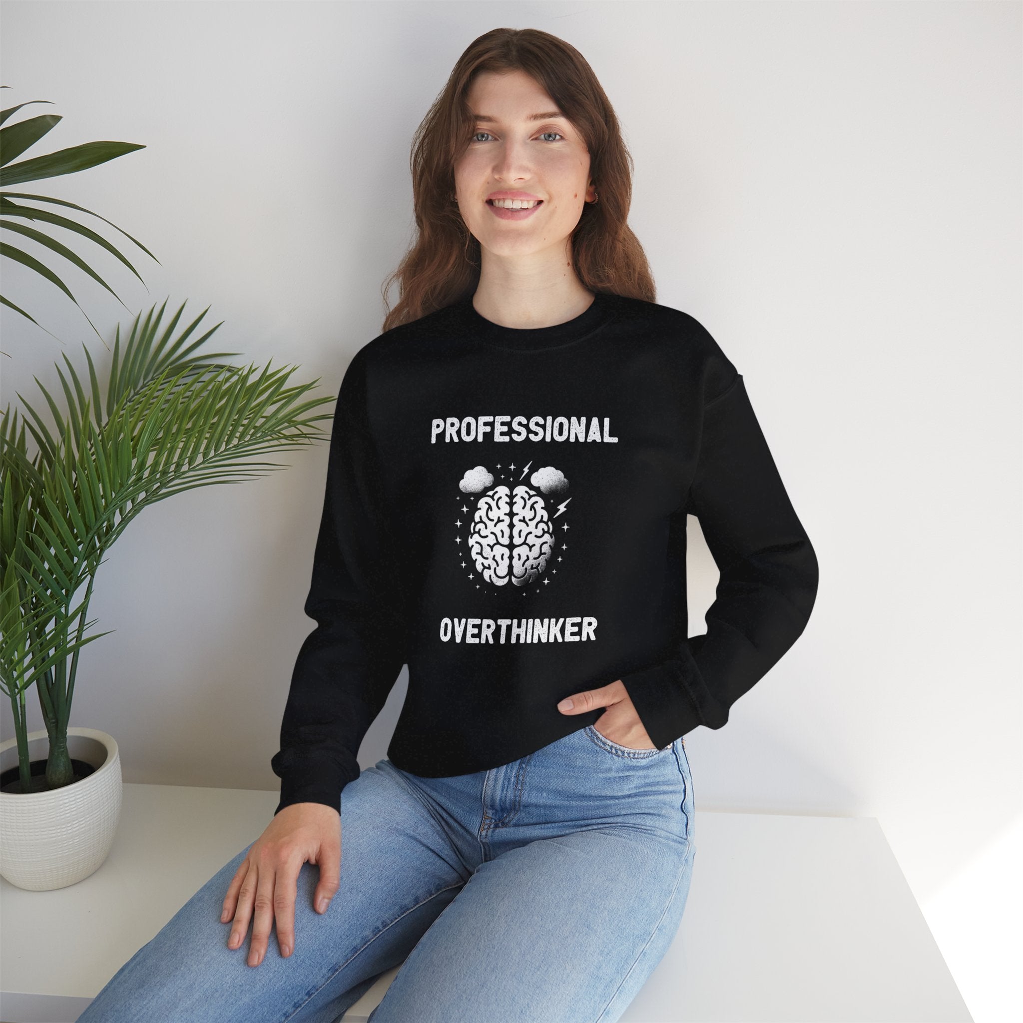 A smiling person sits comfortably on a white surface, cozily wrapped in the "Professional Overthinker - Sweatshirt," adorned with a brain illustration and the words "Professional Overthinker." To the left, a potted plant adds a touch of nature's calm.