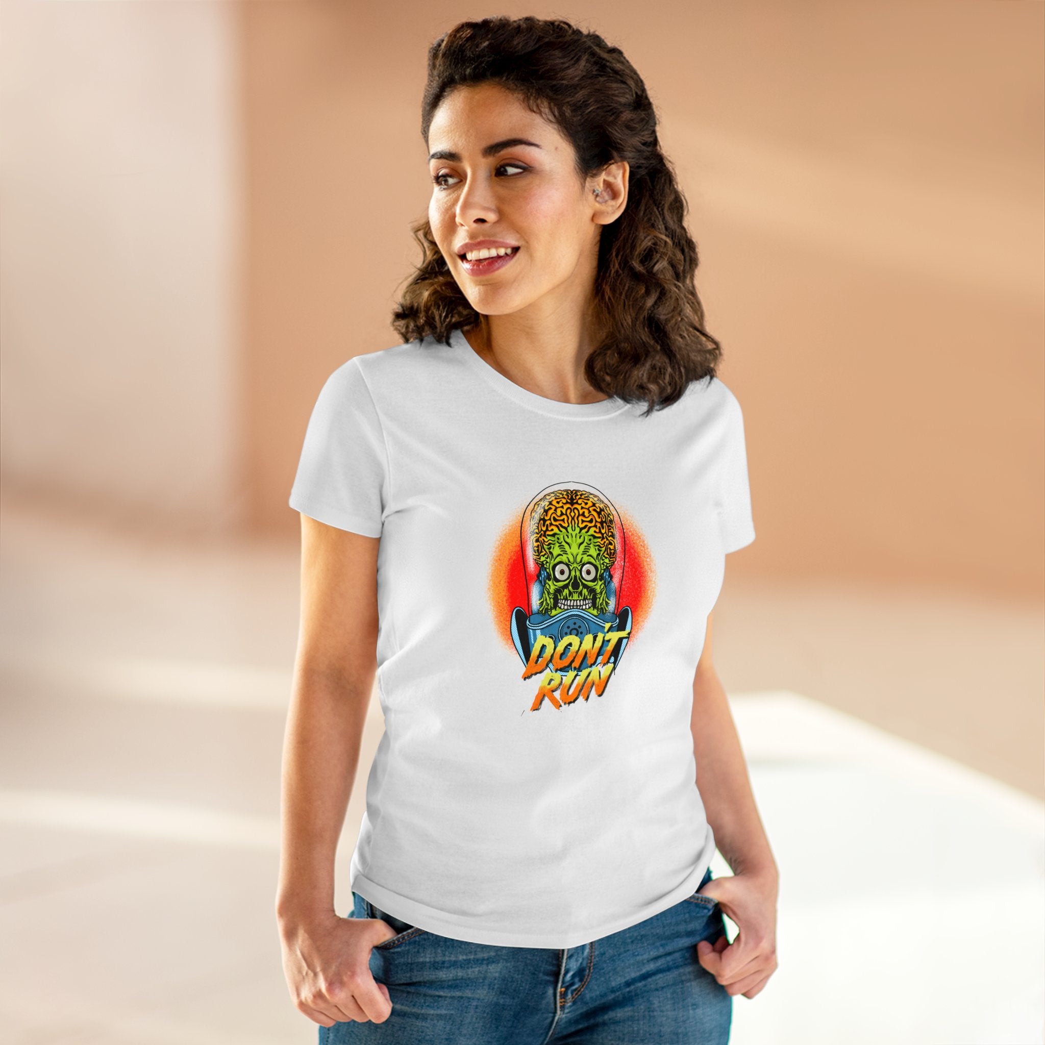 A woman with dark hair wearing the Don't Run - Women's Tee, a white t-shirt made of soft cotton featuring a graphic of an alien head and the text "Don't Run," stands in a well-lit indoor space. The shirt's great fit makes her look effortlessly stylish.