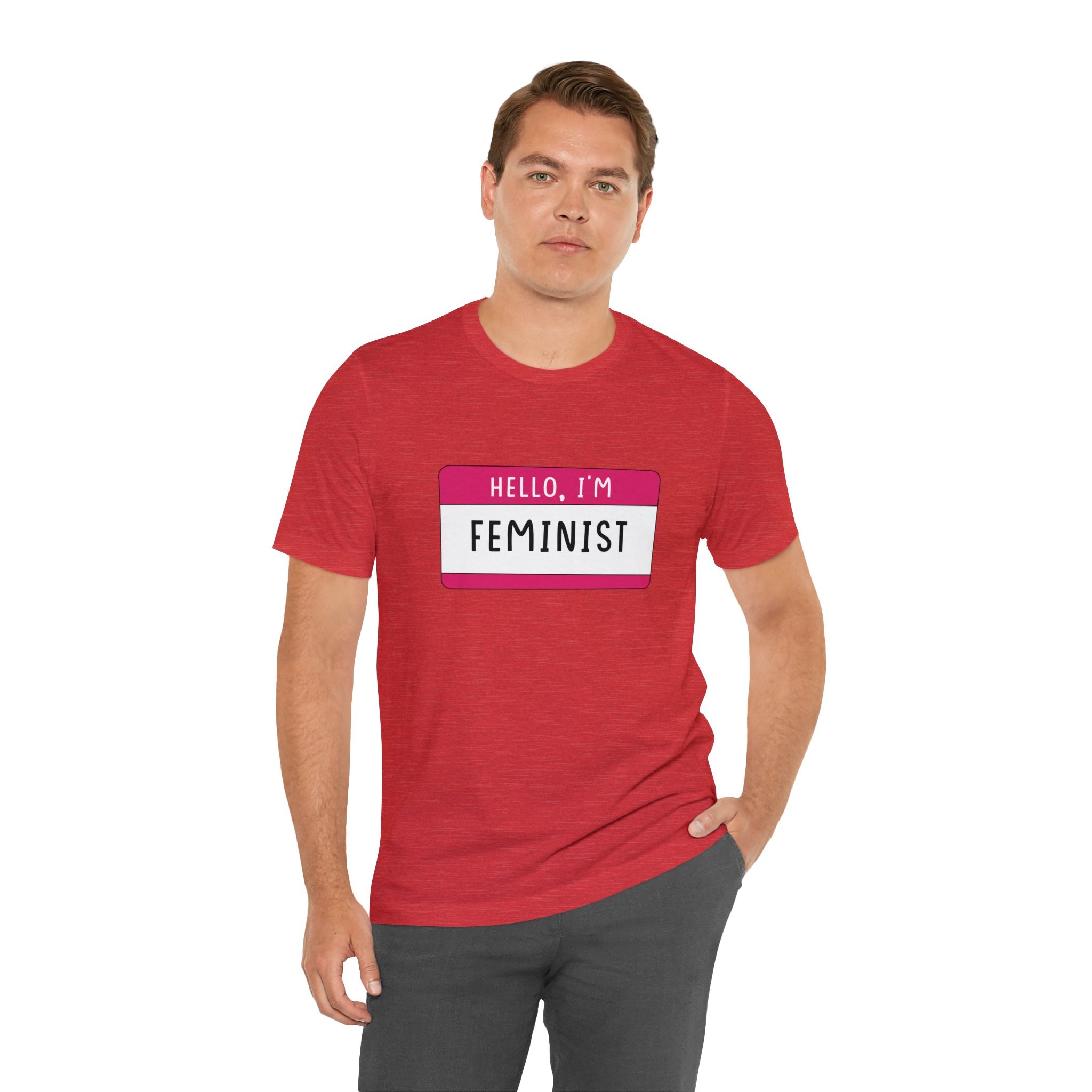 Man in a red "Hello, I'm Feminist T-Shirt" with an "equality" name tag, standing with one hand lightly resting on his hip, against a white background.
