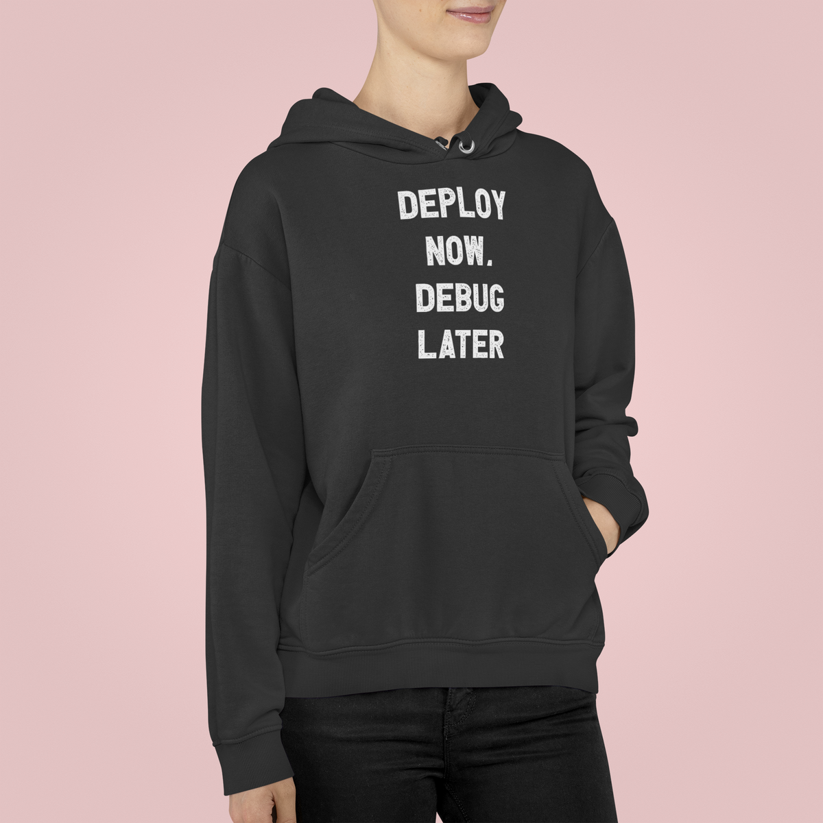 'Deploy Now, Debug Later' hoodie in black, woman wearing it against pink background, funny developer gift