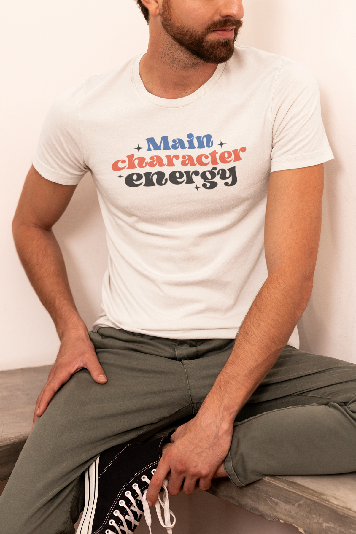 'Main Character Energy' t-shirt in cream, man sitting outdoors wearing it