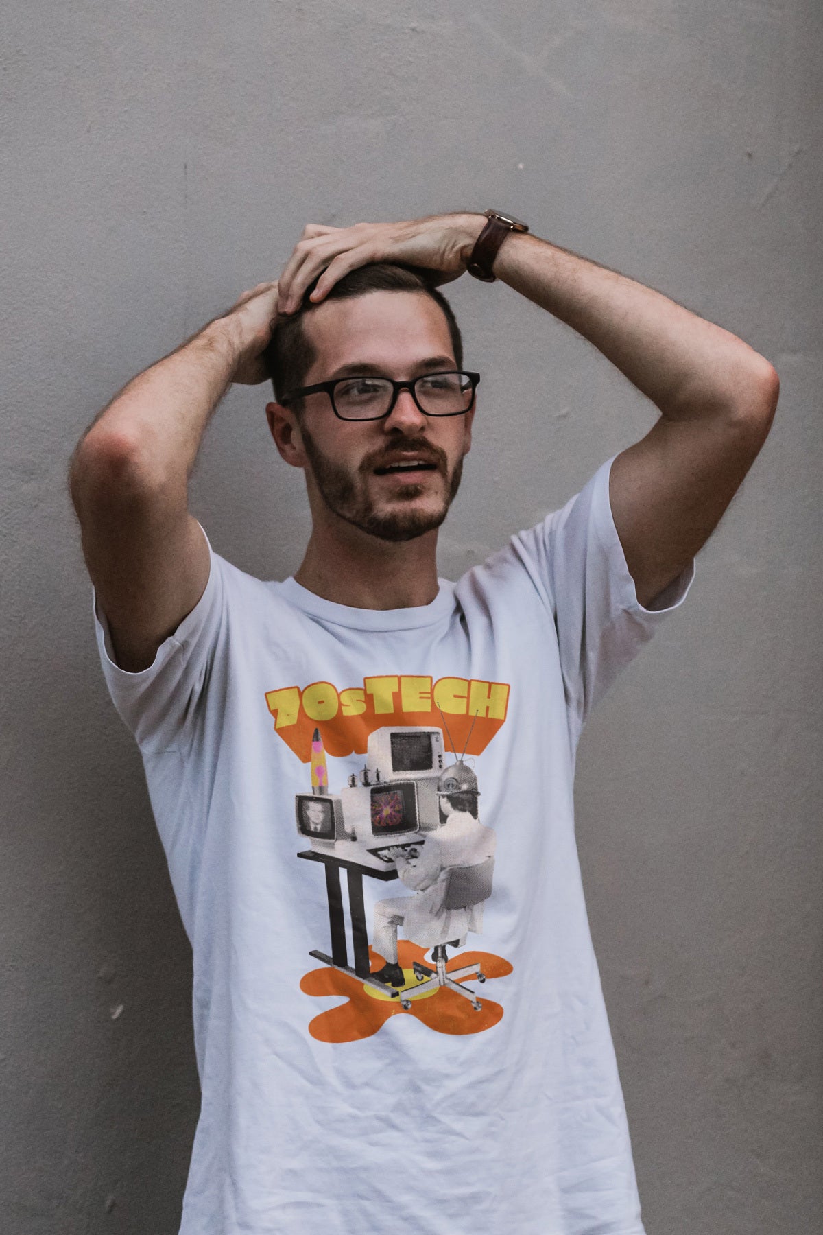 A man wearing 70s Tech glasses and a t - shirt leaning against a wall.