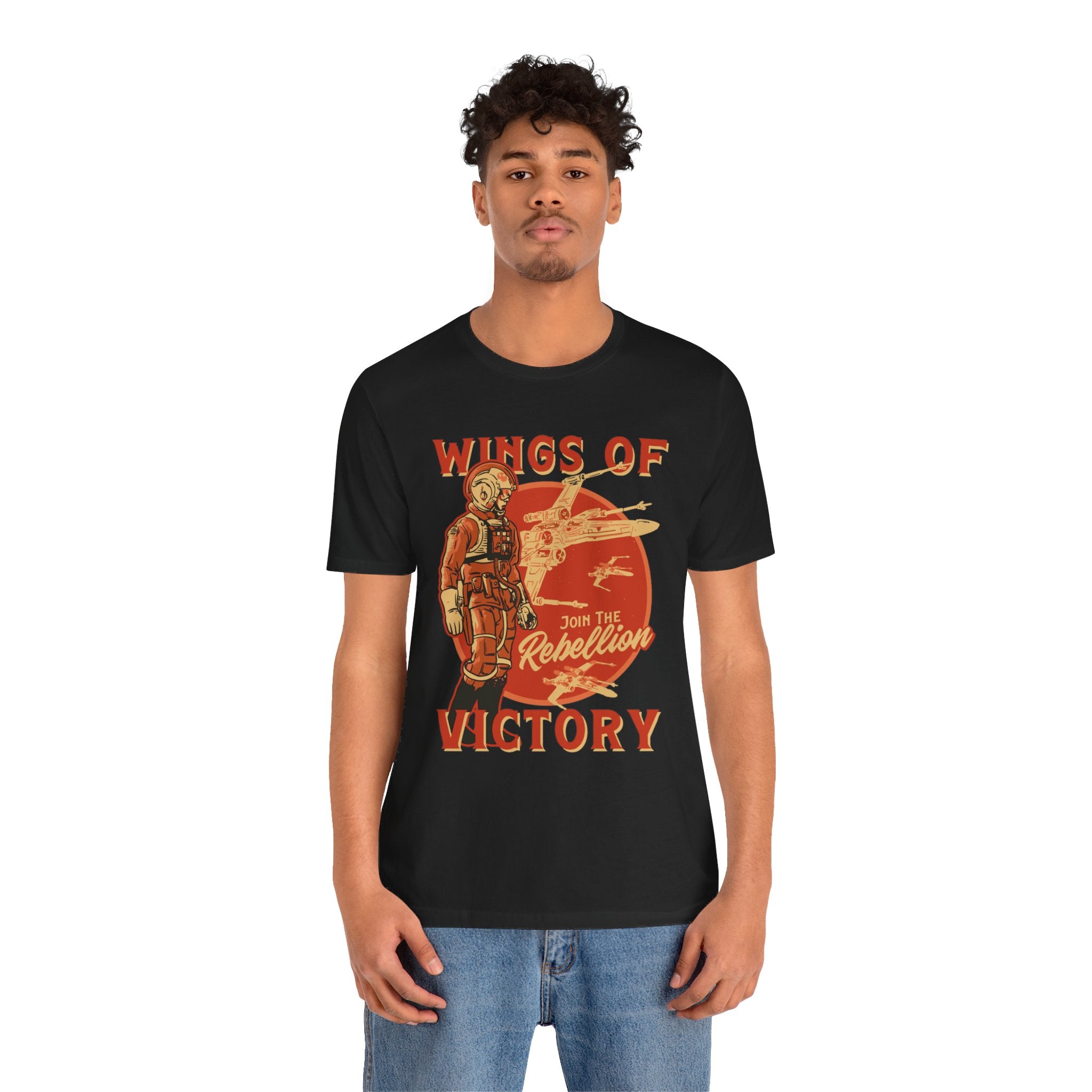 A man stands wearing a "Wings of Victory" T-shirt, a classic unisex jersey tee in black. It features the text "Wings of Victory" and "Join the Rebellion" alongside a vintage-style graphic. Made from soft cotton, this high-quality design complements his short curly hair and blue jeans perfectly.