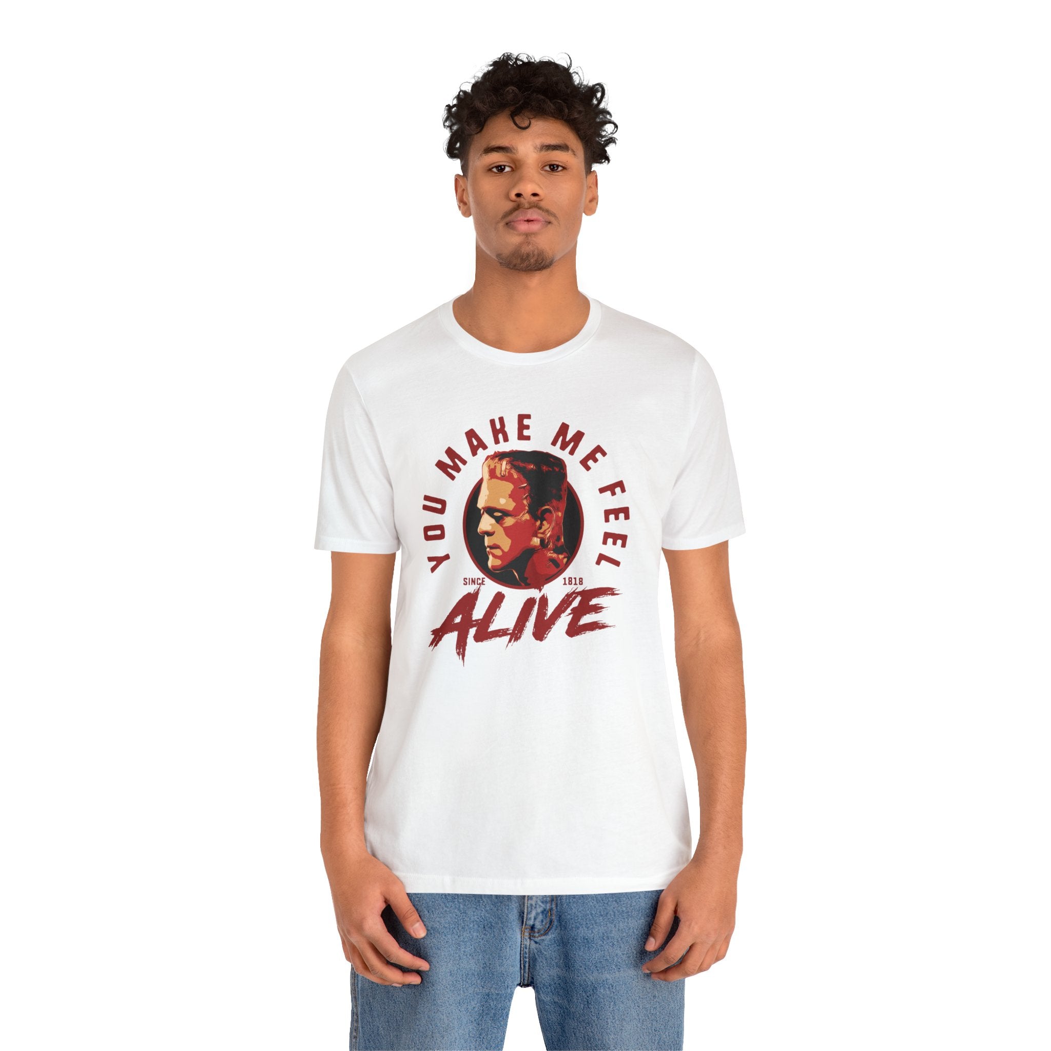 A young man wearing a white unisex You Make Me Feel Alive tee with a quality print and blue jeans stands against a white background.